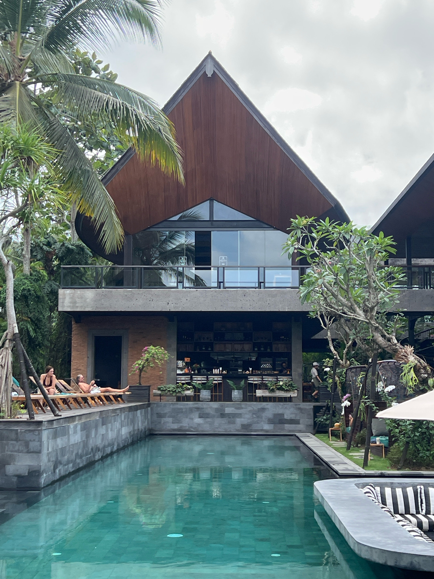 A resort with a swimming pool and lounge chairs is pictured. The resort is located in the Ubud District of Indonesia. The resort's architecture features modern design with wood and glass elements. The swimming pool is surrounded by a stone patio and lush vegetation. Two people are seen relaxing on lounge chairs by the pool. The sky is overcast.