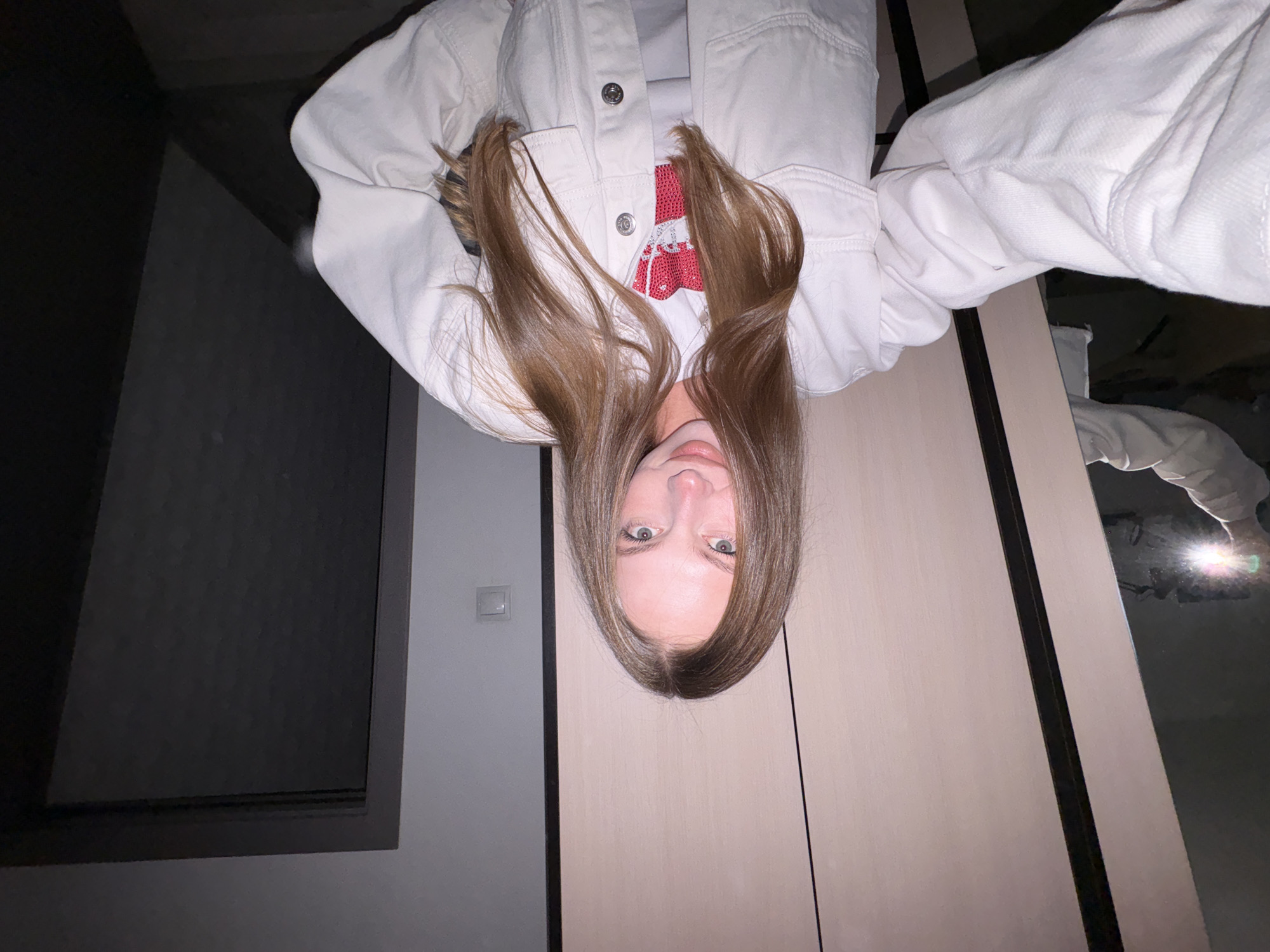 A young female subject is depicted in a selfie, reportedly taken in Perm, Russia. The image is rotated 90 degrees clockwise, showing the subject's head and upper torso from a slightly low perspective as she looks directly at the camera with a subtle, soft smile. She has long, straight, brown hair with a central parting, cascading over her shoulders and partially obscuring the left side of her face. Her eyes are light green or blue-green, and her lips are naturally pink and slightly parted. She is wearing a white denim jacket with silver snap buttons, under which a garment with a red and white pattern is visible.
The indoor background features light grey walls. In the upper left portion of the frame, a dark grey rectangular element, possibly a window frame or recessed architectural feature, is visible against the wall. Below this, a white rectangular light switch or electrical outlet is mounted on the light grey wall. The right side of the frame is dominated by a light beige or pale pink paneled surface, likely a wardrobe or closet door, featuring thin dark horizontal lines indicating panel divisions. In the lower right corner of the image, a bright reflection of a mobile phone's flash is visible, confirming the selfie method, along with a partial, distorted reflection of the subject's hand, arm, and the white jacket sleeve. The scene is illuminated by artificial light, with the phone's flash creating a distinct glare.
