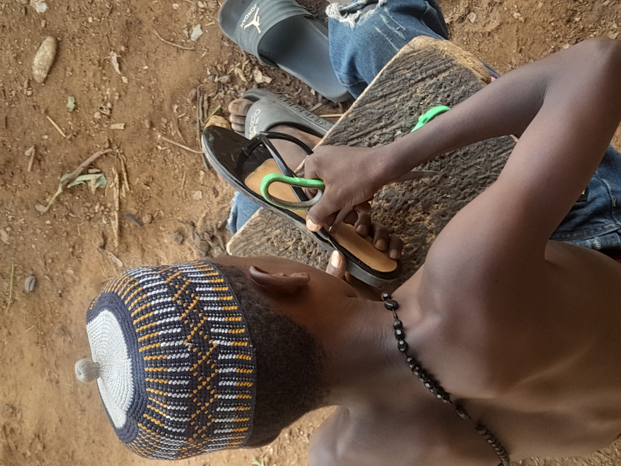 A high-angle shot depicts a person in Akwanga, Nigeria, likely outdoors on a dirt surface. The individual, wearing blue jeans, Jordan and Adidas sandals, sits with their legs crossed on a rough-hewn wooden surface. They wear a knit cap with a white top and a repeating black, white, and gold pattern along the sides, and a beaded necklace. The person is holding a black sandal with a green strap. The surrounding environment is earthen.