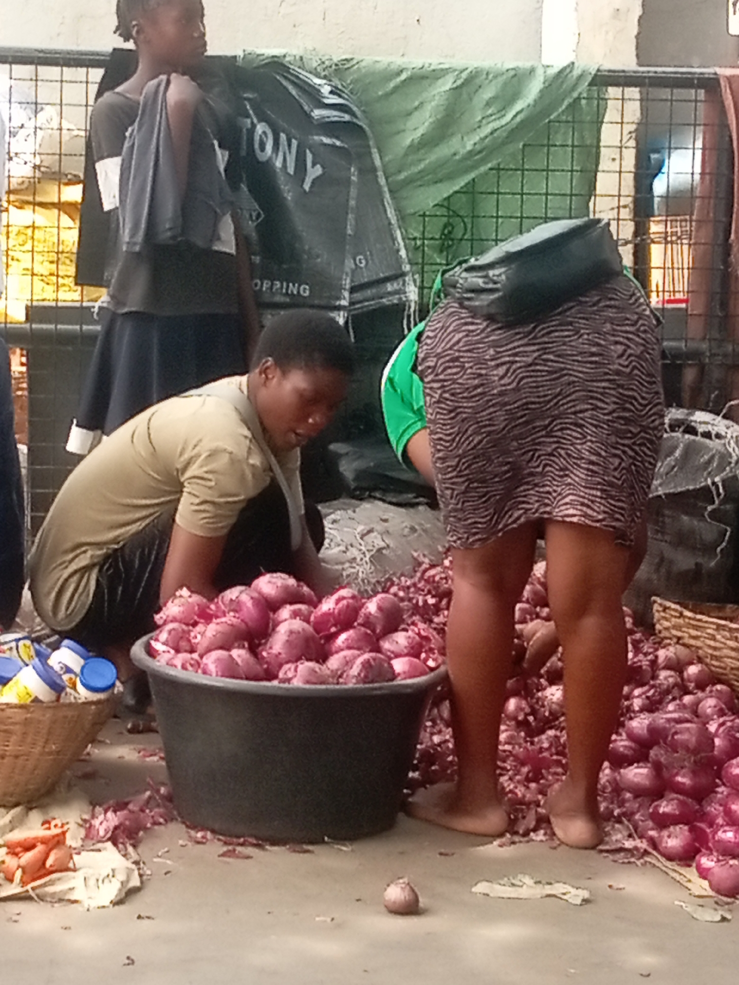 A street scene in Onitsha, Nigeria, shows three people, one crouching and two standing near a large pile of red onions. A woman is crouched next to a large black bucket overflowing with onions, sorting through them. Another woman stands behind her, wearing a green shirt, a zebra-striped skirt, and a black purse. To the left, a young girl stands holding a gray cloth on her shoulder. Various containers are scattered around. A black sack labeled "TONY SHOPPING BAG" is behind the people, along with a section of green mesh.