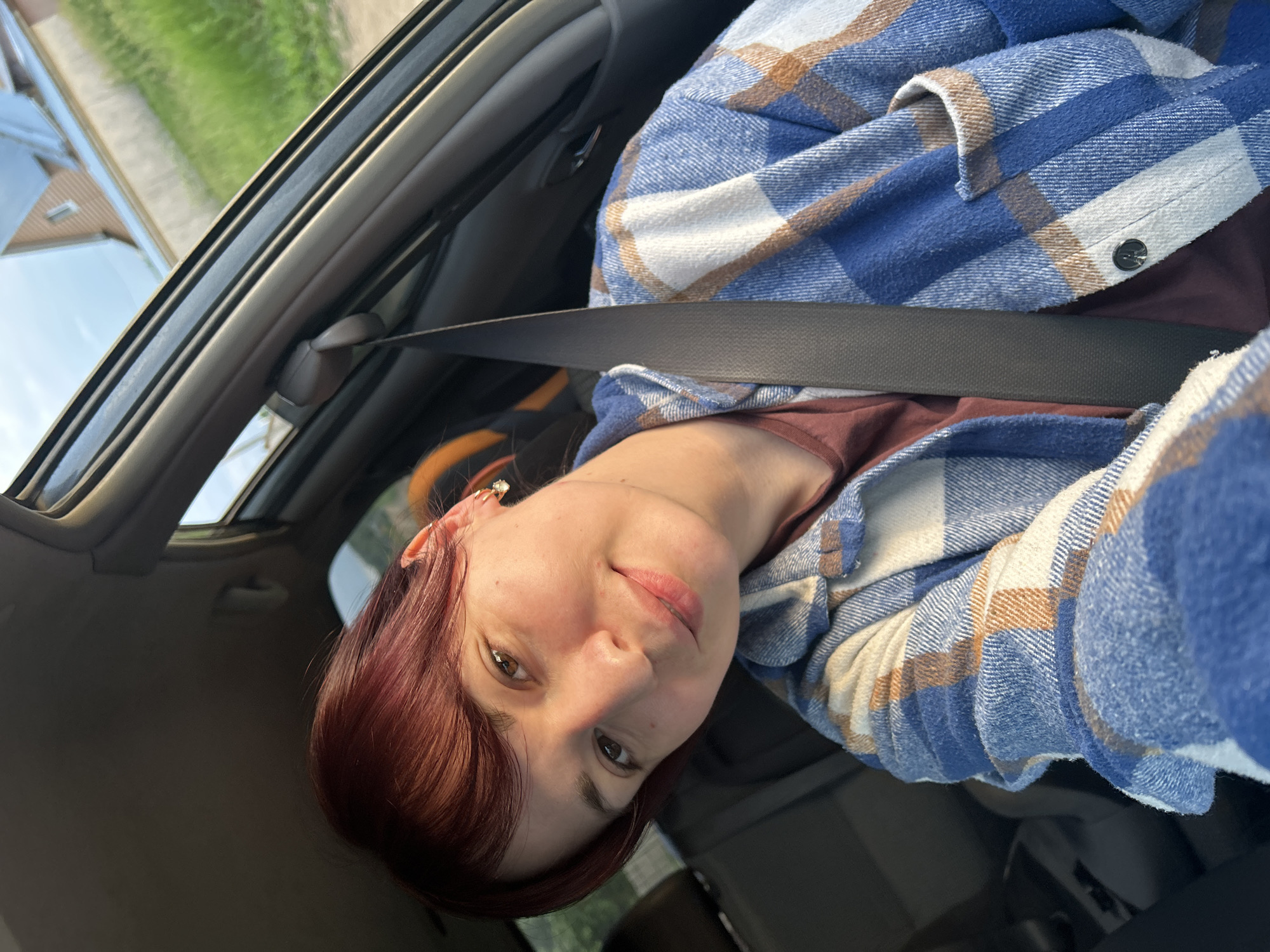 The image shows a young woman with reddish-brown hair and light skin, wearing a blue and white plaid shirt over a maroon t-shirt. She is seated in the passenger seat of a car, with a seatbelt fastened across her chest. She is looking at the camera and smiling, with her head slightly tilted. Her right ear is visible and has a dangling earring with a clear stone. The interior of the car, including the dashboard and a portion of the passenger seat, is visible. Through the car window, a blurry background shows green grass, a light-colored building with a blue roof, and a clear sky, suggesting an outdoor setting. The lighting indicates it is daytime, possibly late afternoon due to the warm tones. The scene is set in the city of Fokino, Russia.