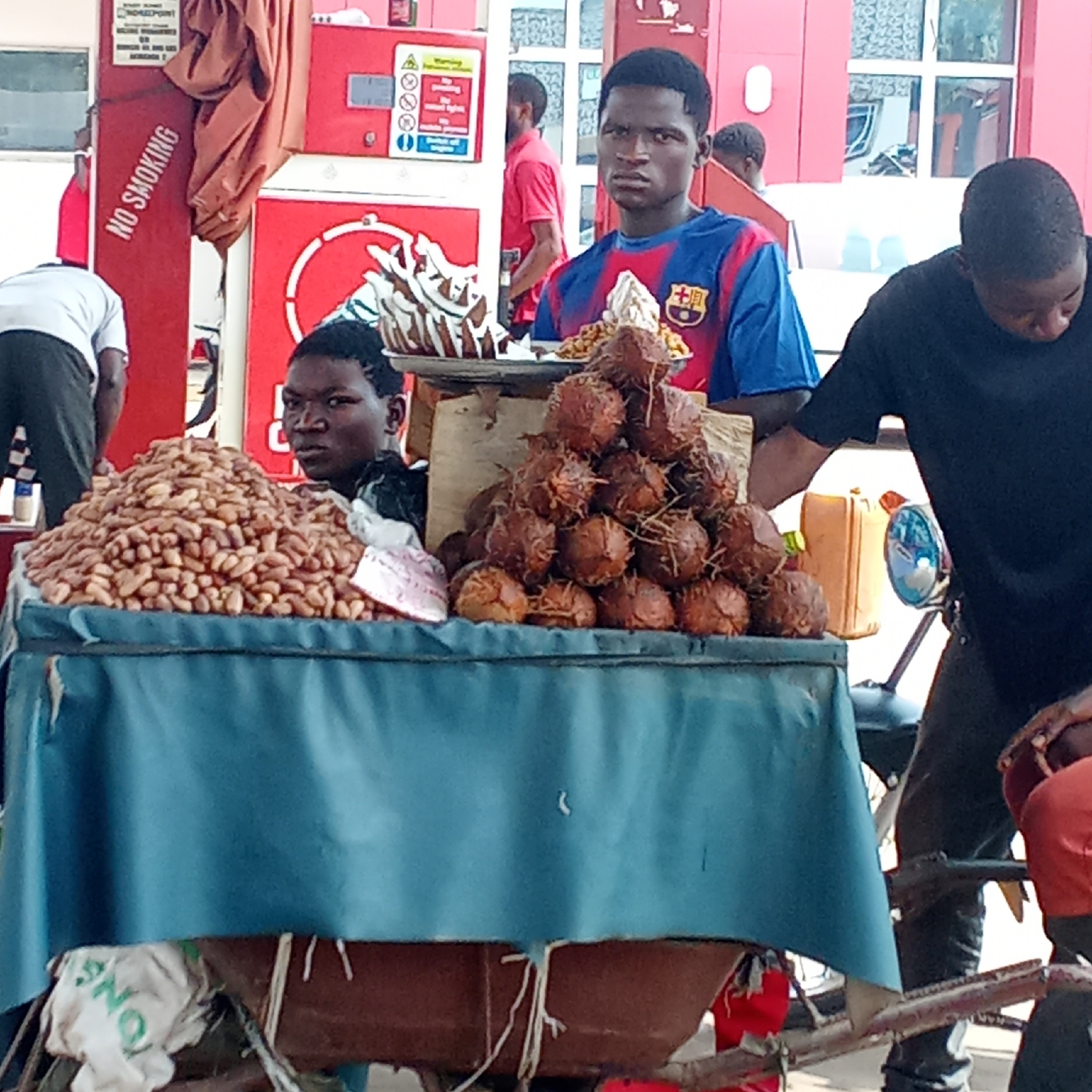 A street vendor's table in Akwanga, Nigeria, displays a large pile of peanuts on the left and a pyramid of brown, spiky-skinned fruits, likely coconuts, on the right. Two young men stand behind the table, one wearing a blue and red FC Barcelona jersey and holding a tray with additional food items. To the right of the vendor is a man dressed in a black T-shirt who is standing near a motorcycle. The vendor's table is covered with a blue cloth, and a "No Smoking" sign is visible in the background, along with gas pumps.
