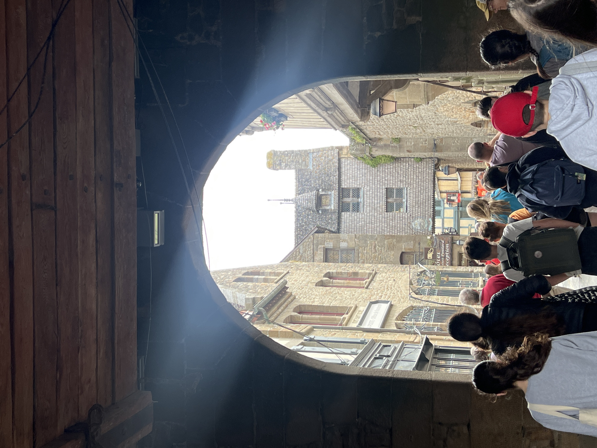 A low-angle shot looks through a stone archway towards a narrow street lined with old stone buildings. A group of tourists walk away from the camera into the street. The buildings have steeply pitched roofs, small windows, and are constructed of rough-hewn stone. The sky is bright and overcast. The wooden beams of the archway are visible overhead. A few businesses are indicated by signs, one appearing to be "LA POSTE - Office de Tourisme." The scene appears to be in Le Mont-Saint-Michel, France, a tidal island commune known for its medieval architecture and abbey.
