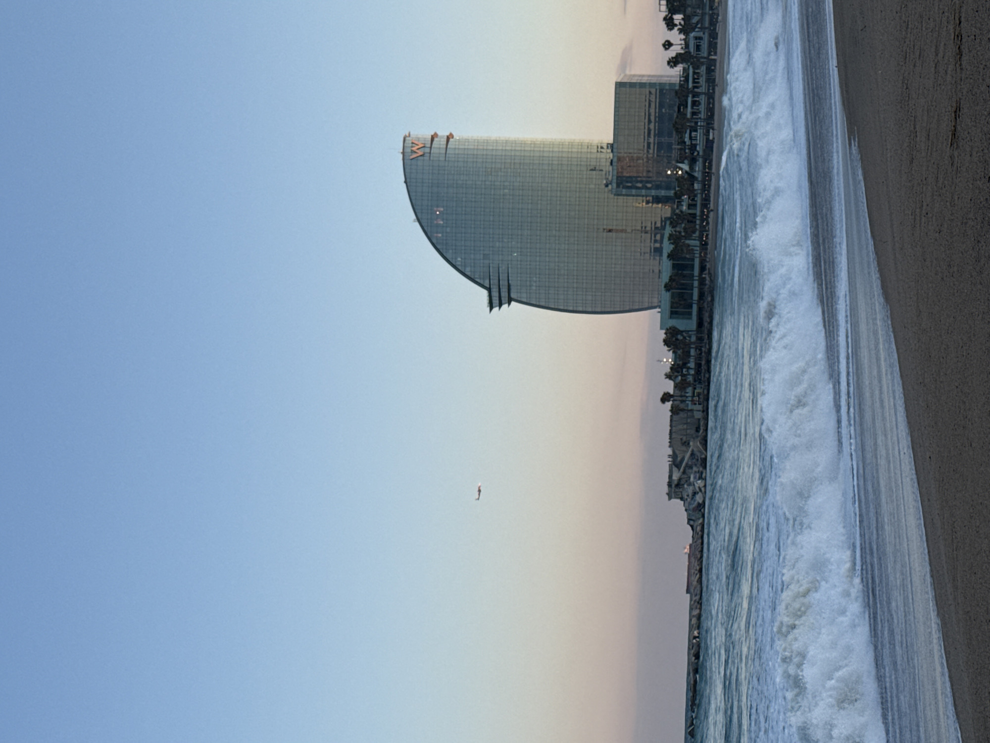 This image shows the W Barcelona hotel on Barceloneta Beach in Barcelona, Spain. The beach is on the right side of the image. The sky is on the left. A small aircraft is in the sky between the hotel and the horizon.