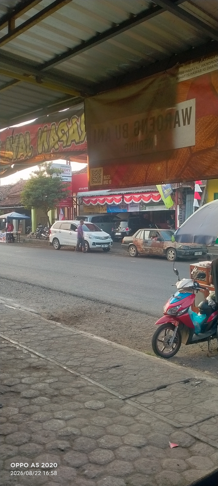 The image captures a street scene in Kedung, Indonesia, featuring a roadside establishment with a large banner advertising "AYAM NAGAN" and "WAROENG BU...". The banner is partially obscured by a metal roof structure supported by yellow beams. In the foreground, a red and blue scooter is parked on the side of a paved road. Further down the road, a white SUV is parked, and a person in a purple shirt is standing next to it. To the right of the SUV, a rusty, light-brown sedan is also parked. The environment appears to be an urban or semi-urban setting, with buildings visible behind the parked vehicles and trees lining the street. The paved ground in the foreground is a combination of hexagonal and rectangular blocks. The timestamp "OPPO A5 2020 2026/08/22 16:58" is visible in the bottom left corner.