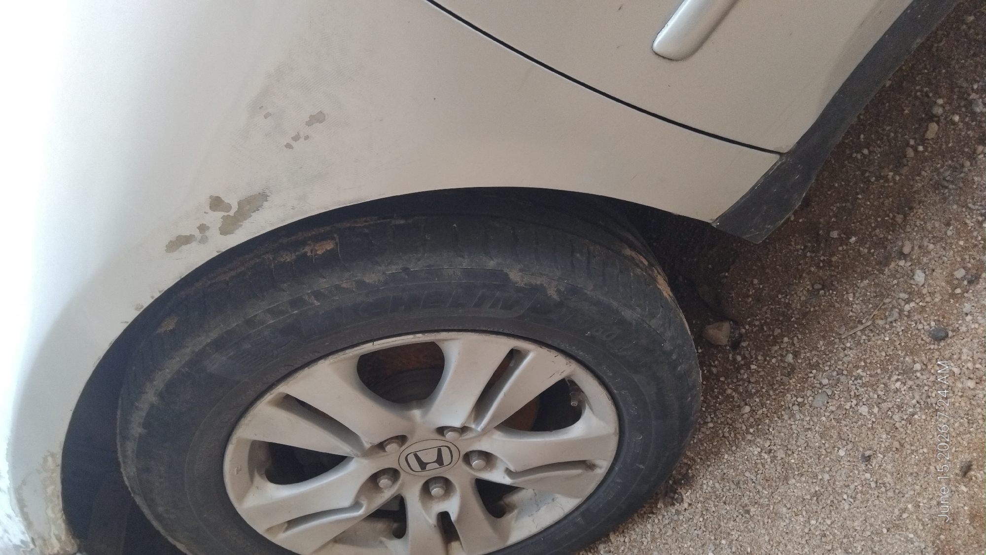 A close-up, low-angle shot shows the front passenger-side wheel and fender of a light-colored sedan. The tire is black and appears worn, with dirt and debris on its surface. The silver alloy wheel has five spokes, and a Honda logo is visible in the center. The fender has several patches of missing paint and primer, revealing exposed metal, and some dirt smudges. The car is parked on a surface of small gravel and dirt. The timestamp "June 15, 2026 7:24 AM" is overlaid in the bottom left corner of the image. There are no people or distinct environmental features that would definitively place the scene in Howayi, Nigeria, but the arid ground cover is consistent with a dry, possibly rural or unpaved area in Nigeria.