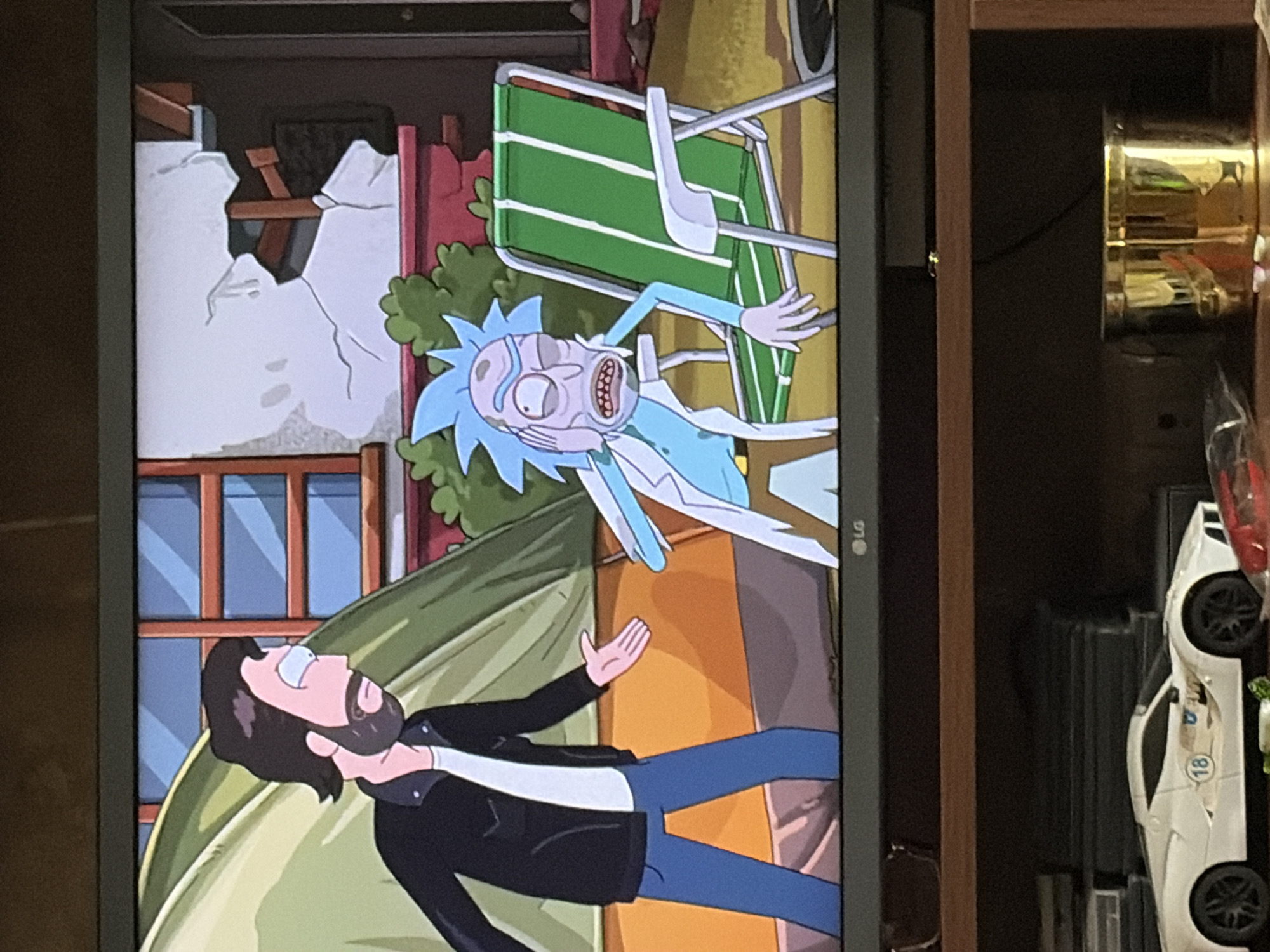 The image displays a television screen showing an animated scene from the show Rick and Morty. On the left is a male character with dark hair and a beard, wearing a black jacket, white t-shirt, and blue pants, standing with his arms outstretched. To his right is Rick Sanchez, characterized by his blue hair and lab coat, sitting in a green and white striped lawn chair. The background depicts a damaged building with exposed wooden beams and cracked white plaster, alongside a green tent and lush green foliage. Below the television, on a wooden shelf, a white toy sports car with the number 18 is visible, along with a gold-colored cylindrical object. The scene is framed by the television screen and the wooden shelf.