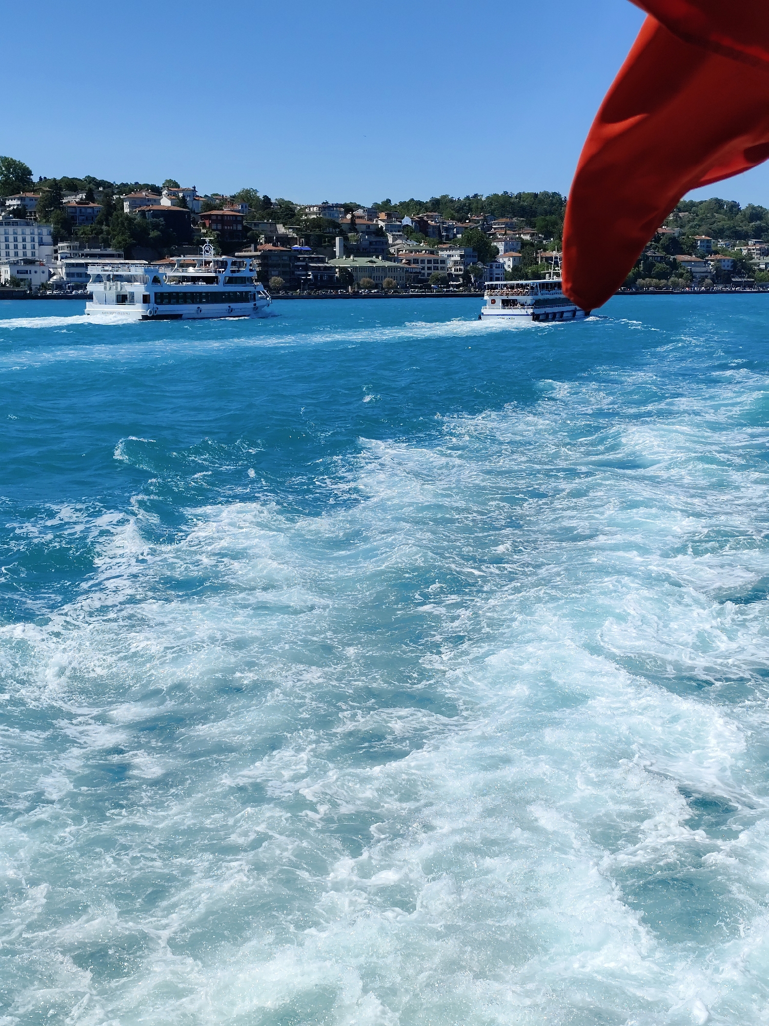 The image captures a marine scene in Sarıyer, Turkey, with two passenger ferries navigating the turquoise waters. In the background, the shore is dotted with buildings and verdant trees. The view is from a moving vessel, evidenced by the swirling wake that dominates the foreground. An indistinct orange object, possibly a flag, is visible in the upper right corner, partially obscuring the scene. The sky is clear and blue.