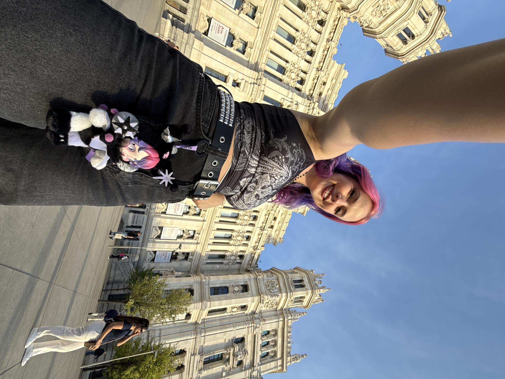 A selfie shot shows a woman smiling and looking at the camera, with her arm extended. The woman has dyed hair of pink and purple, a black tank top with a silver design, and a studded belt. A doll and other charms are attached to her belt. The background reveals a cityscape, specifically a building facade. The location appears to be in Madrid, Spain, based on the architectural style of the building. People are seen walking in the distance. The sky is blue and clear.