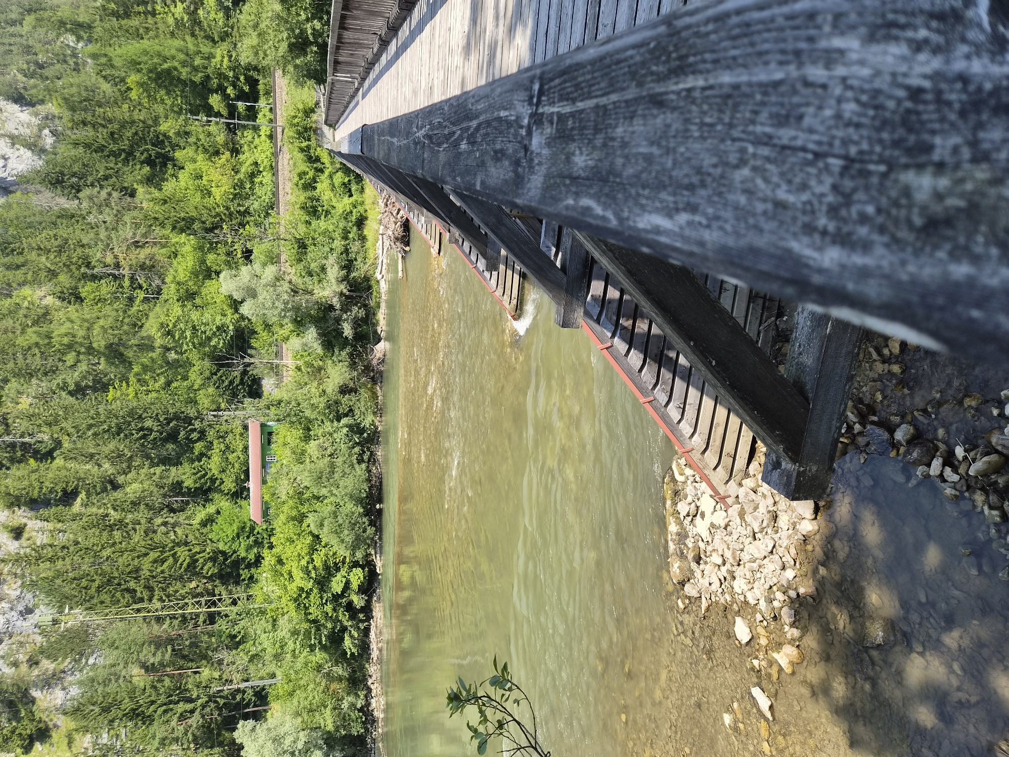 This is a photograph taken from a wooden bridge, looking towards a river and a forested mountainside. The bridge, constructed of weathered wood, extends into the distance, with its railing and planks visible in the foreground and mid-ground. The river, with its greenish-brown water and rocky bed, flows gently. Along the far bank of the river, dense green trees line the base of a steep, rocky mountain. In the mid-ground, nestled amongst the trees, is a small, red-roofed building. A set of train tracks is visible above the building, running parallel to the river. The overall scene depicts a natural, somewhat remote environment, likely in the vicinity of Admont, Austria, known for its mountainous terrain and the Admont Abbey. There are no people visible in the image.