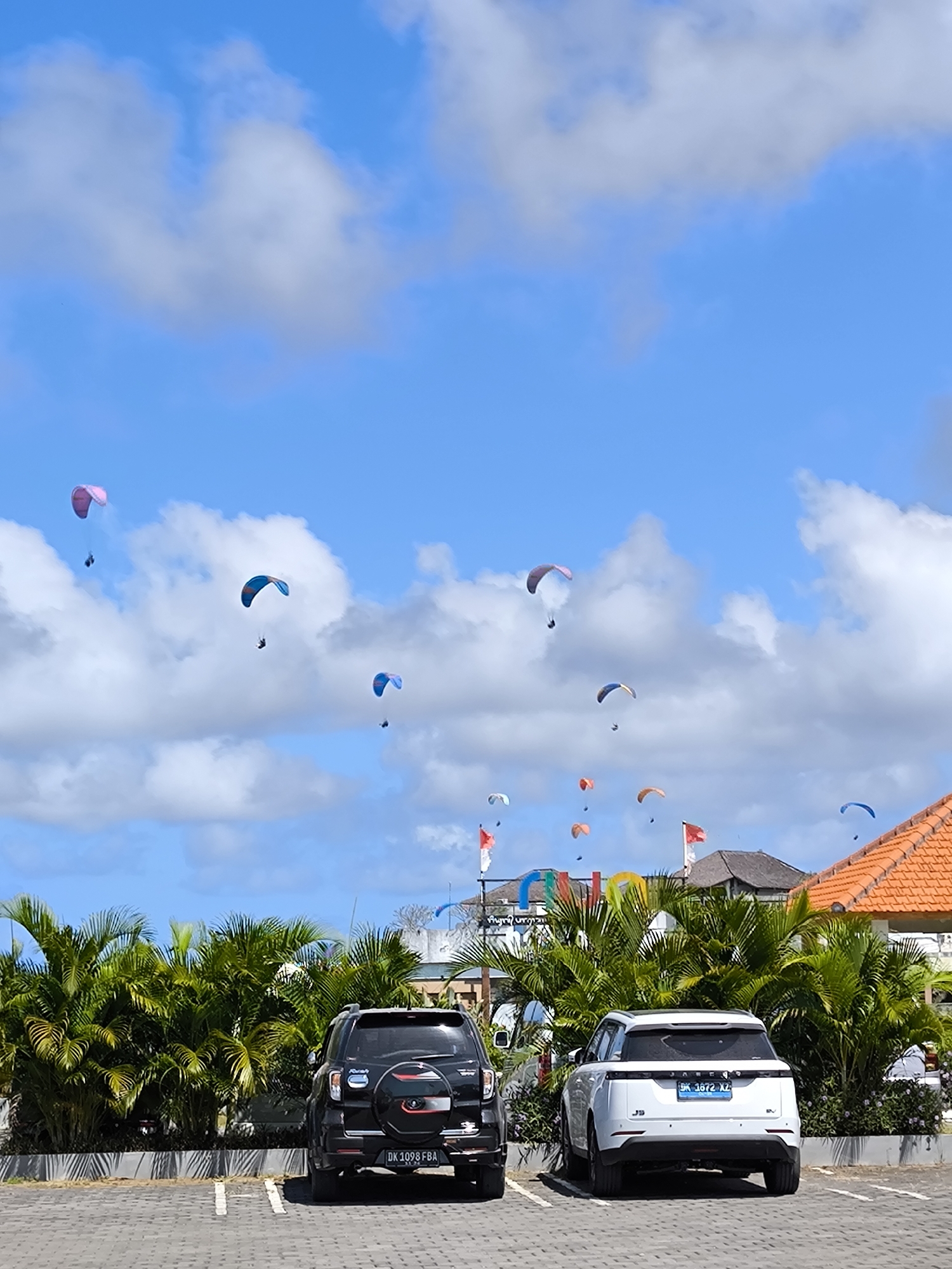 The image captures a parking lot scene in an unknown city, featuring two parked cars, a black SUV, and a white SUV. The registration plate of the black SUV reads "DK 1098 FBA", while the white SUV displays "DK 1672 XZ". Behind the cars, there's a row of lush green palm trees, partially obscuring the view of a building with a distinctive orange tiled roof. In the sky above the building, multiple paragliders are airborne, dotting the blue sky with their colorful canopies. The sky is partly cloudy.