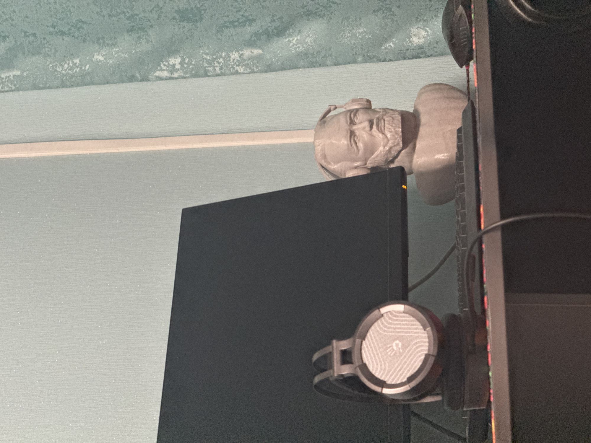 A medium shot shows two computer monitors and a statue bust against a light blue wall in an indoor setting. A gray statue bust depicting a bearded man is positioned on top of the monitor to the right, its gaze oriented to the right. Gray and white headphones are attached to the side of a computer monitor. The setting appears to be a room or office space, but there is no location information.