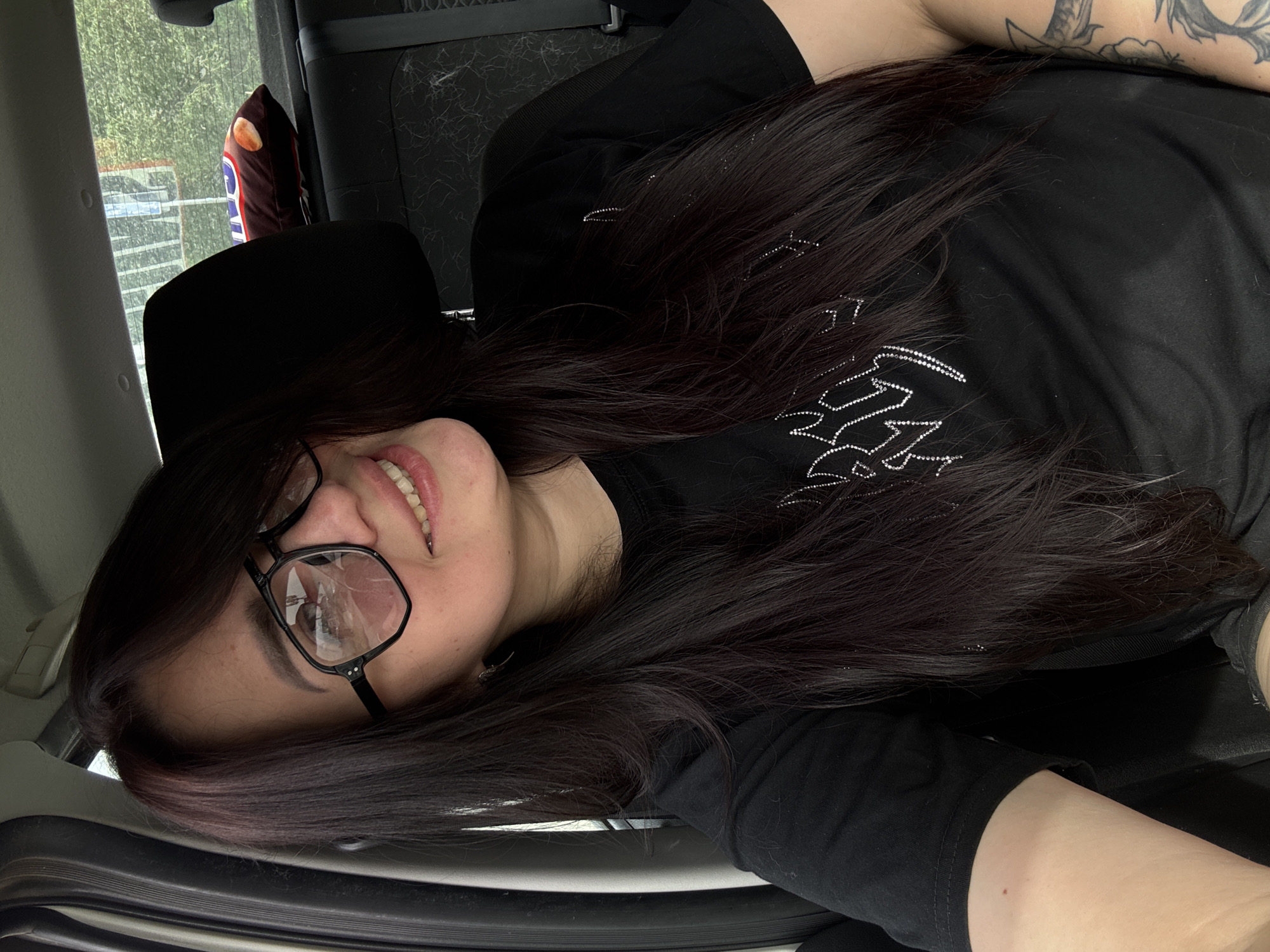 A woman with long, dark hair and glasses is lying down in what appears to be a car. She is wearing a black shirt with a design on it. Part of her arm, with a tattoo, is visible. A headrest and part of a bag of chips are visible in the background. It's difficult to determine the exact location.