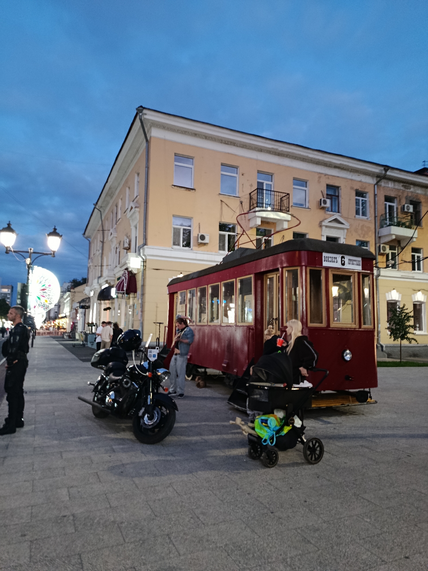 The image captures a street scene in Saratov, Russia, at dusk. In the foreground, a dark motorcycle is parked on a paved walkway. To the right of the motorcycle is a red vintage tram with wooden trim. Two women are interacting near the tram, one holding a red cup and the other looking into a stroller. A man is playing a violin next to the tram. Further down the street, several people are walking, and on the left, a man in dark uniform stands near a lamppost. A Ferris wheel is visible in the distance, illuminated against the twilight sky. The buildings lining the street have yellow facades and balconies. The overall atmosphere suggests a public space with some form of entertainment or gathering.