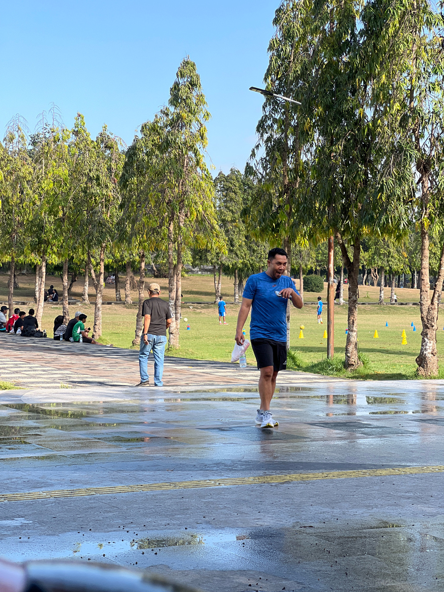 The image depicts a park in Denpasar, Indonesia, with a focus on a man in a blue t-shirt and black shorts walking towards the camera on a wet, paved pathway. He is smiling and holding a white object in his right hand, and a plastic bag in his left. The pathway is bordered by trees with lush green foliage and a grassy area beyond, dotted with yellow and blue cones, suggesting an outdoor activity or training session. In the background, several people are seen sitting and standing in the shade of the trees, while others are engaged in activities on the grass. The sky is clear and blue, indicating a sunny day.