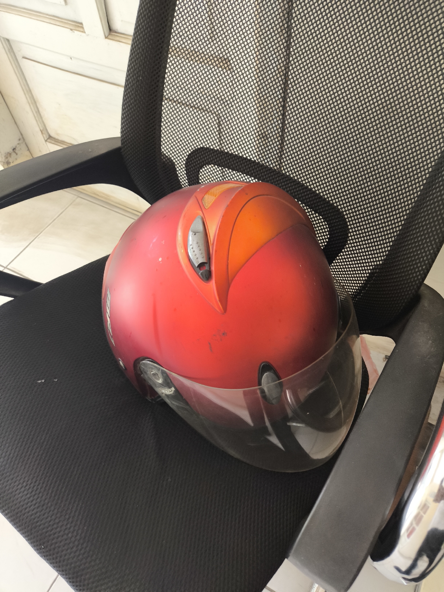 A red helmet with a clear visor rests on the black mesh seat of an office chair. The chair is positioned in front of a white-paneled door. The scene is indoors, possibly in a home or office, and no people are visible. The floor appears to be tiled. The lighting suggests it is daytime. The image quality is adequate, with some minor dust particles visible on the helmet and chair.