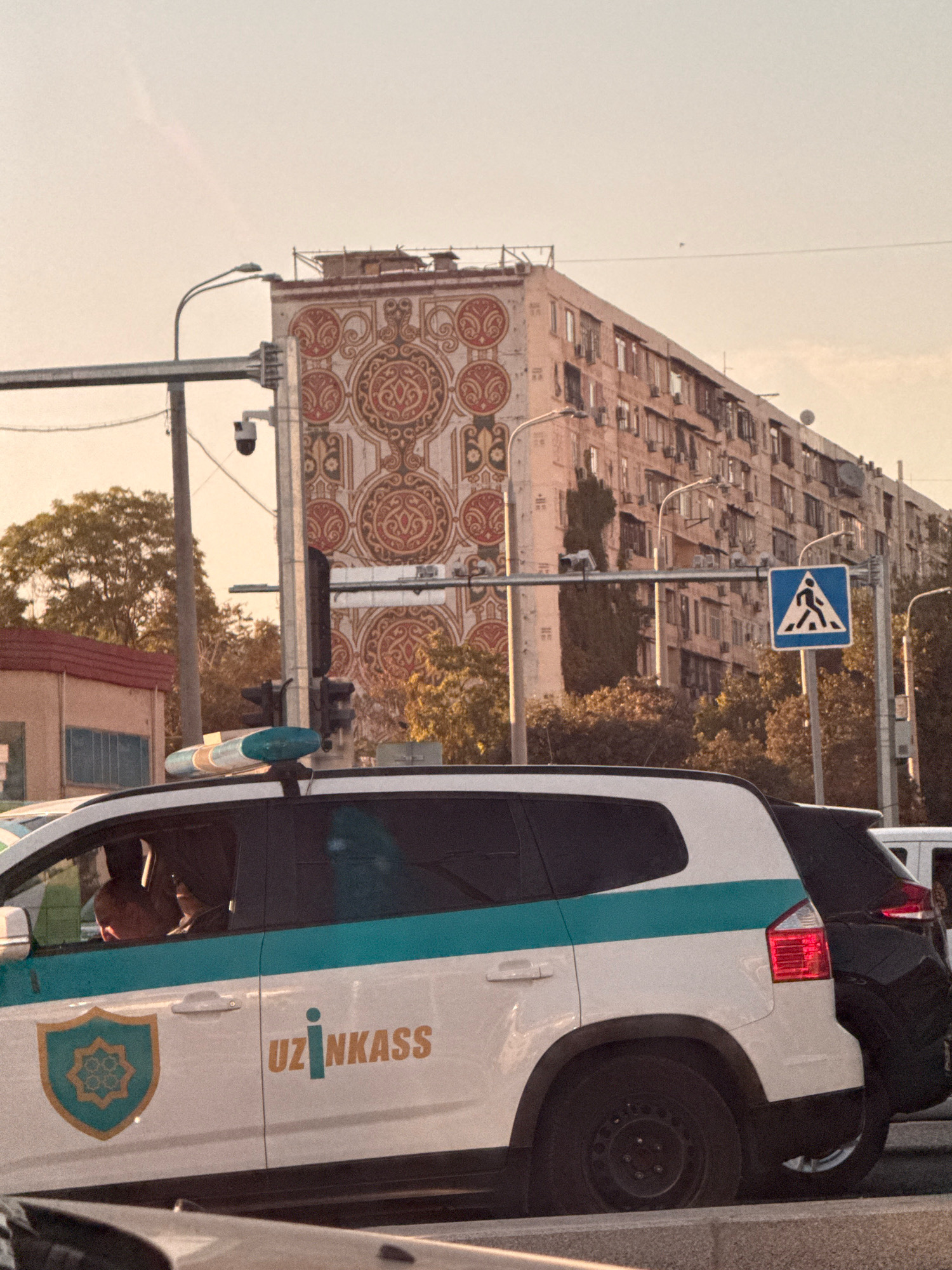 A street scene in Tashkent, Uzbekistan, features an "UZINKASS" branded vehicle, likely a security or transport service, with a driver visible. Behind it, a pedestrian crossing sign and an imposing apartment building with intricate decorative panels are prominent. Streetlights, traffic signals, and security cameras are also present. The sky is a pale, hazy orange, suggesting a sunset or sunrise.