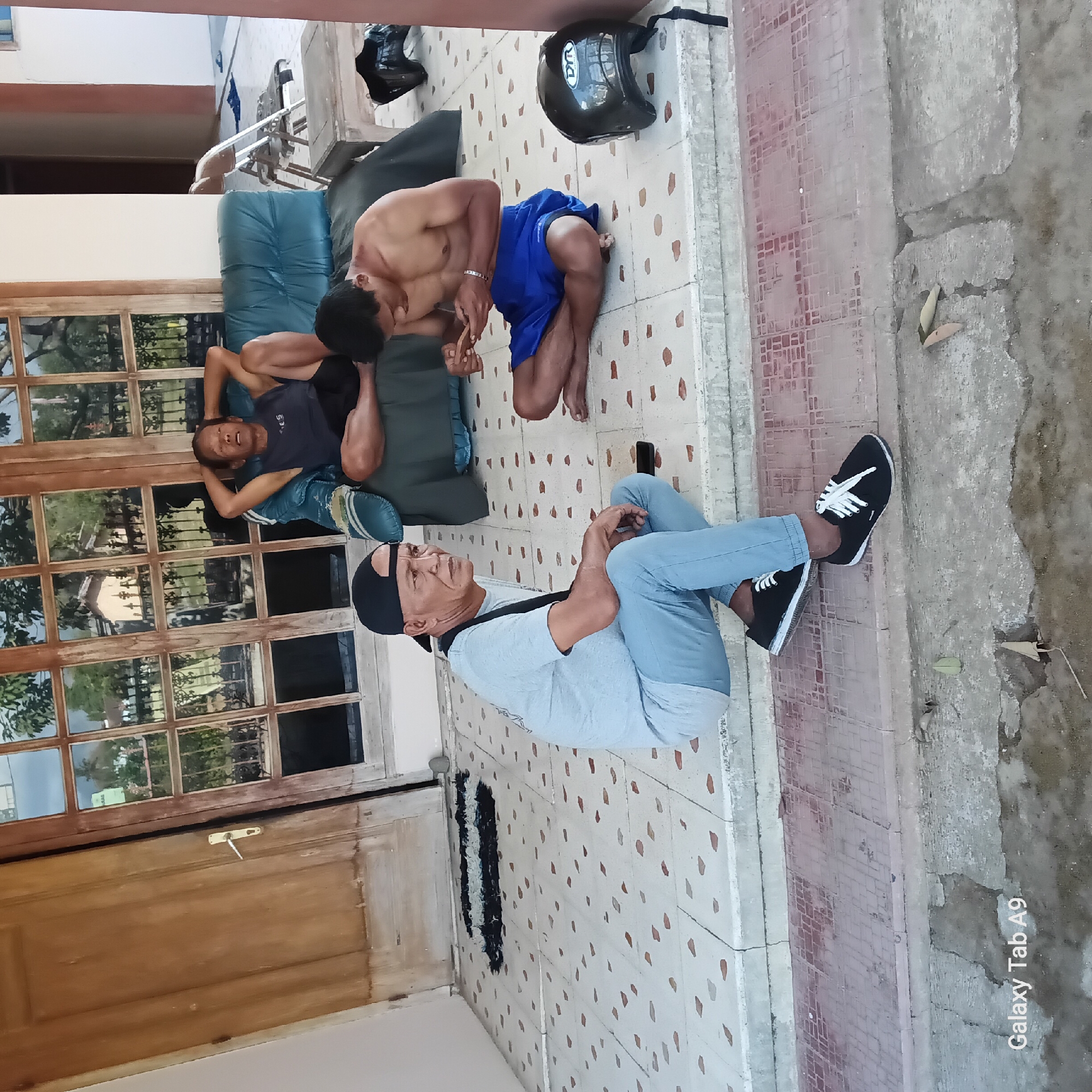 The image shows three men in an outdoor, shaded area, likely a porch or veranda, in what appears to be a residential setting in Sambirejo, Indonesia. The man in the foreground, wearing a black baseball cap, light blue t-shirt, and light blue jeans, sits on the edge of a tiled step, facing left. He has a dark strap across his shoulder and is looking off to the side. To his right, a shirtless man wearing blue shorts sits cross-legged on the tiled floor, engrossed in his smartphone. In the background, another man reclines on a dark green sofa, his hands behind his head, also looking away from the camera. Behind the sofa, a large wooden-framed window with multiple panes reflects the outdoors, showing trees and a building. To the right of the sofa, a partially visible folding chair and a small wooden table are present. A black helmet with a white logo is on the floor near the shirtless man. The floor is tiled with a pattern of light squares and smaller reddish-brown or orange accents. The overall impression is a casual, relaxed gathering outdoors.