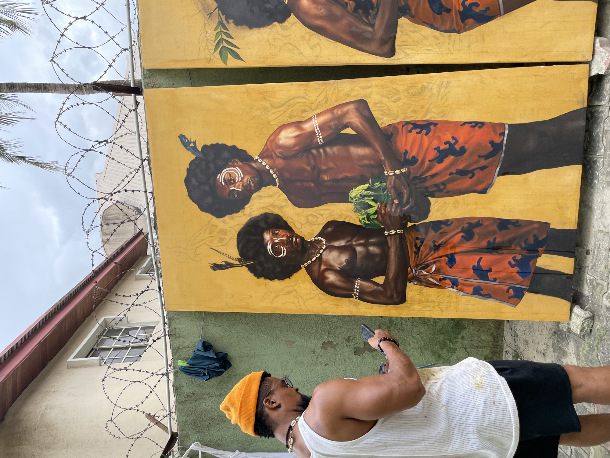 In the city of Ado, Nigeria, a Black man wearing an orange beanie, glasses, a white tank top, and black shorts is standing in front of two large, unfinished paintings. The paintings depict Black men with afros, wearing traditional attire. The man in the foreground is holding a smartphone and appears to be taking a photo of the paintings. The background features a wall with barbed wire on top, a building with windows, and palm trees. The ground is paved with grey stones, and there are some scattered rocks and debris. The overall scene suggests an outdoor art studio or exhibition space.
