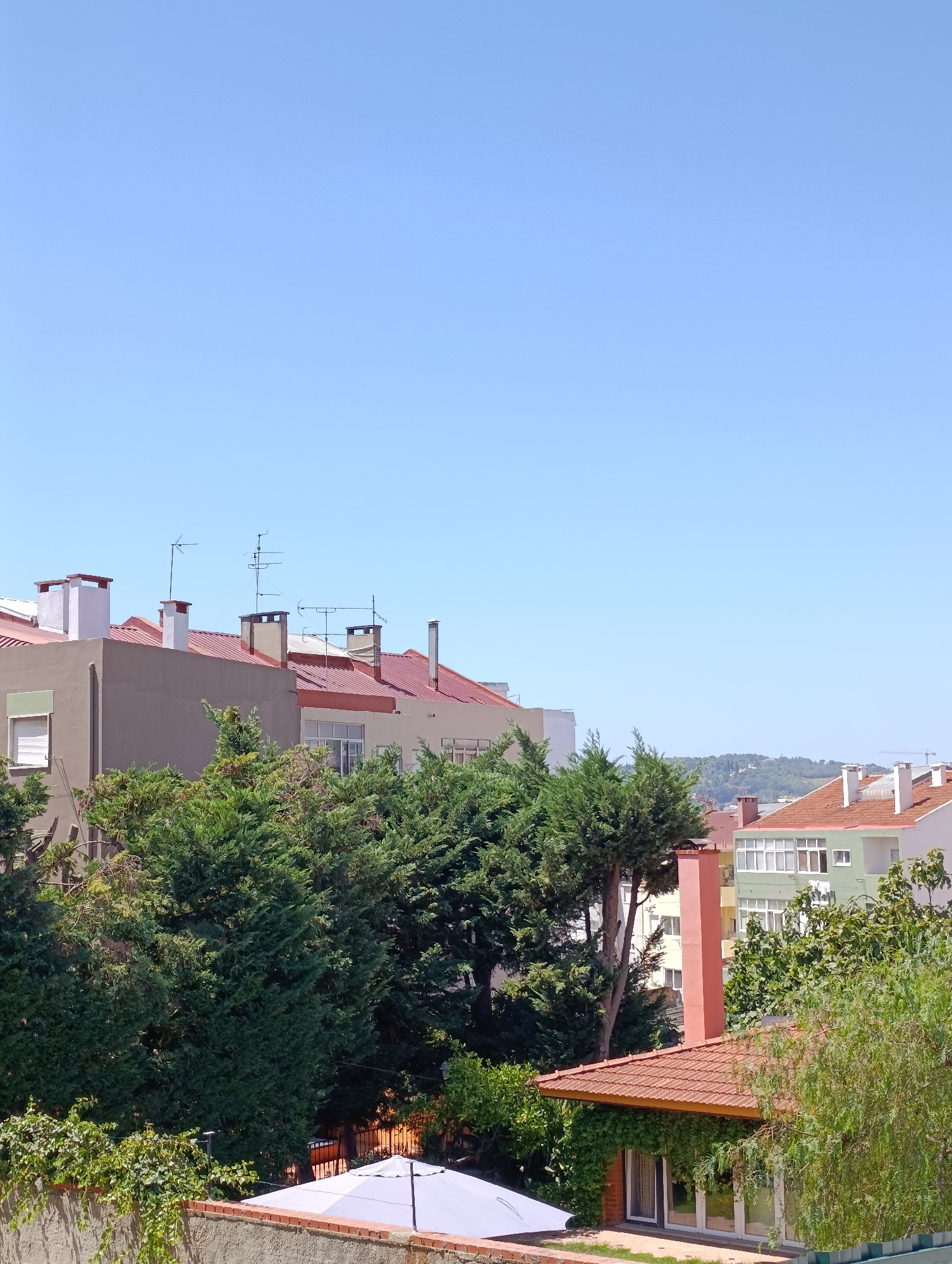 The image shows a cityscape from a slightly elevated vantage point, likely a window or balcony, with a clear blue sky dominating the upper portion. The lower half of the image is filled with rooftops, trees, and architectural elements, suggesting an urban or suburban environment.
In the foreground, a low, textured wall made of concrete and brick is visible, topped with a sparse scattering of green ivy. A portion of a white umbrella with a segmented pattern is situated on the ground behind this wall. Further back, a cluster of dense, dark green coniferous trees forms a significant visual barrier.
Behind the trees, a series of building rooftops are prominent. The closest building features a reddish-brown tiled roof and a salmon-pink chimney. To its left, a two-story building with a reddish-brown tiled roof is visible, characterized by several white chimneys of varying designs, some square and others cylindrical. This building appears to be older, with what look like antique television antennas mounted on the roof. Adjacent to this, another building with a similar reddish-brown tiled roof and white trim extends further to the right. Behind these, other buildings are partially obscured by trees and distance. One building has a light green facade with white-framed windows.
Further in the distance, a rolling, green hillside is visible under the bright sky. The overall impression is one of a sunny day in a developed area with a mix of residential buildings and natural greenery. There are no people or discernible actions taking place. The scene is static and appears to be a snapshot of a typical residential area.