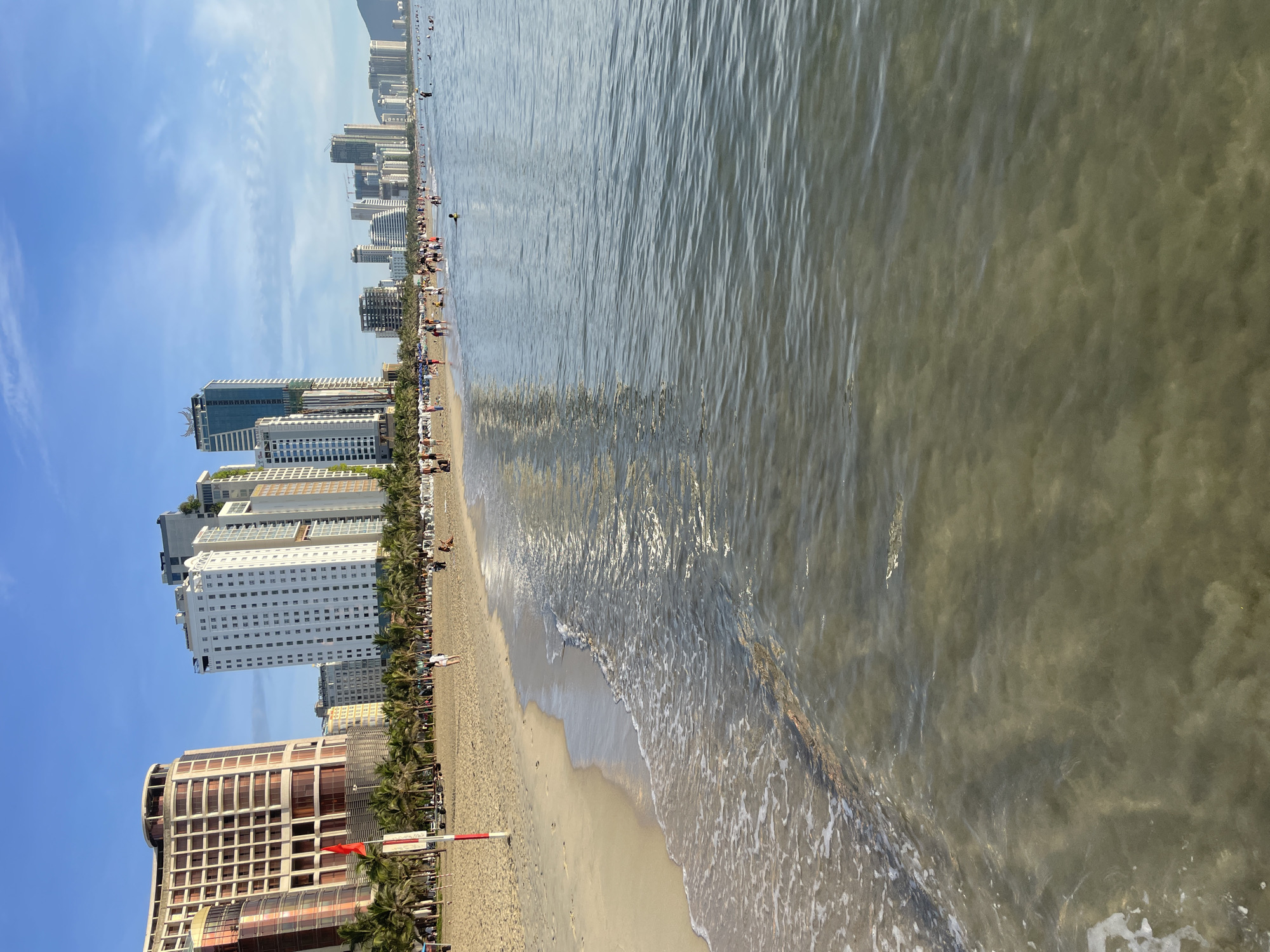 The image depicts a beach scene in Da Nang, Vietnam, characterized by a coastline lined with modern high-rise buildings and a sandy beach populated by numerous individuals. The sea occupies the foreground, with gentle waves lapping at the shore. The beach itself is dotted with palm trees and a multitude of people engaged in various activities, such as walking, relaxing, and swimming in the shallow water. In the background, a cityscape of hotels and residential towers rises above the palm trees, indicating an urban coastal environment. The sky is partly cloudy, suggesting fair weather conditions. The overall impression is that of a bustling tourist destination.