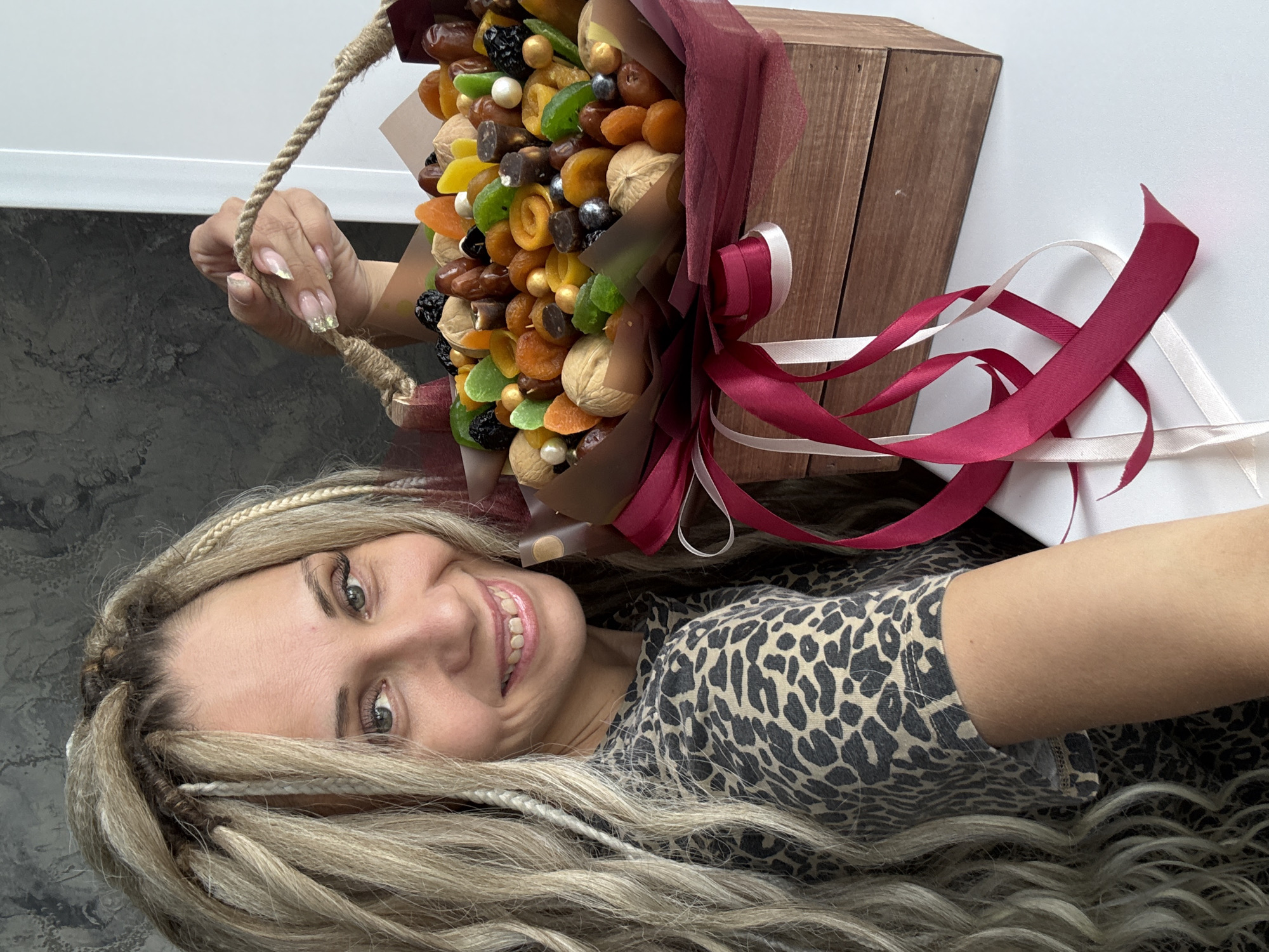 The image depicts a woman with long, blonde hair styled with braids and wearing a leopard-print shirt. She is smiling and holding a bouquet of dried fruits and nuts in a wooden box tied with red and white ribbons. The bouquet contains various dried fruits, including dates, apricots, and green and yellow candied fruits, along with walnuts and decorative gold and silver balls. The background is a white wall, and the woman appears to be indoors. There is no information indicating that this image was taken in Rusanovo, Russia.