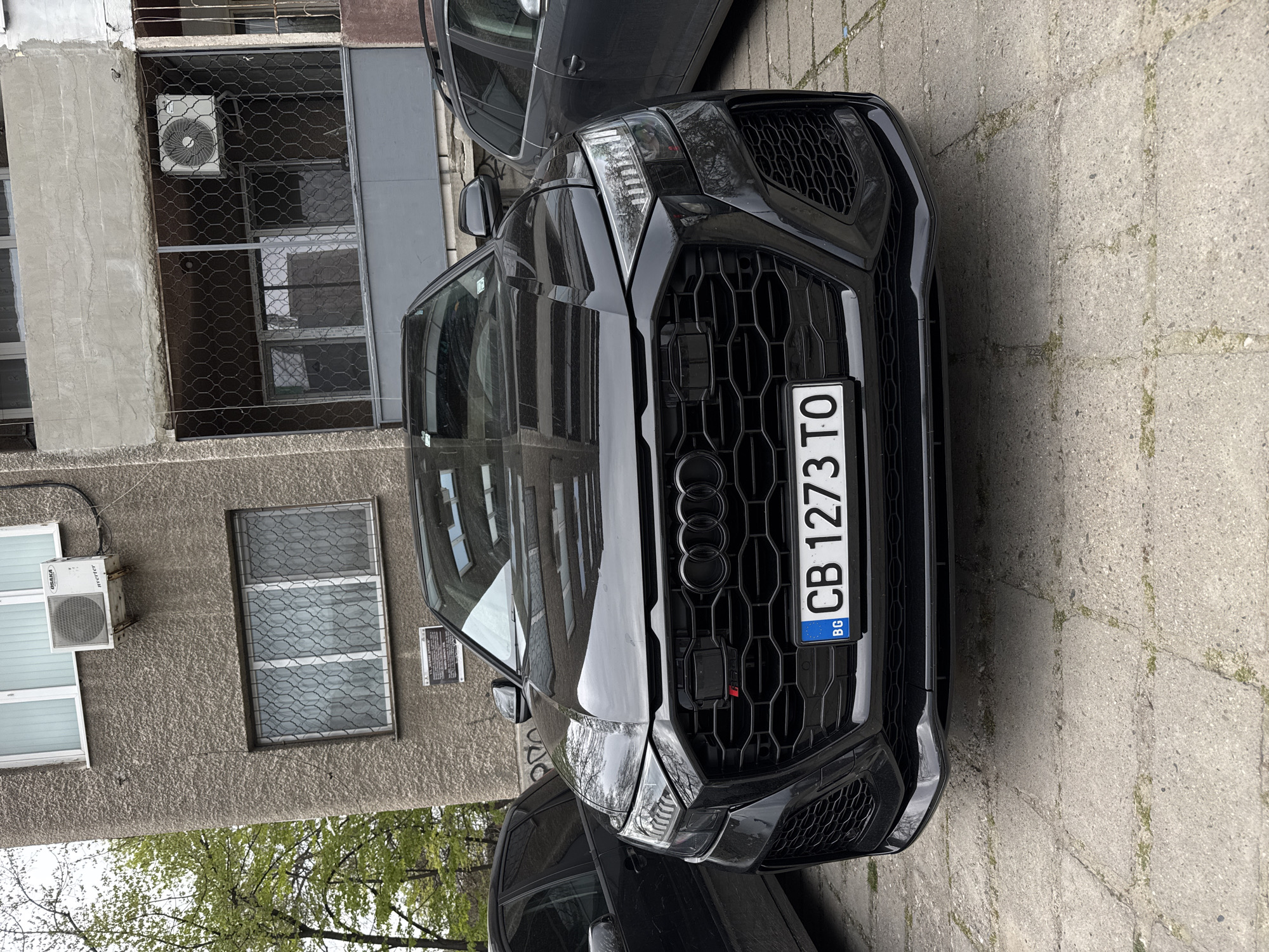 The image shows a black Audi RS Q8 parked on a concrete surface adjacent to a building. The car's front is prominently displayed, featuring its grille, headlights, and license plate, which reads "CB 1273 TO" and indicates registration in Bulgaria ("BG").
The building is gray, with windows protected by wire mesh and external air conditioning units. A portion of another dark-colored vehicle is visible in the background and on the right side of the frame. There are no people visible in the image. The environment appears to be an urban street. Based on the license plate and architectural style, it is likely located in Plovdiv, Bulgaria.