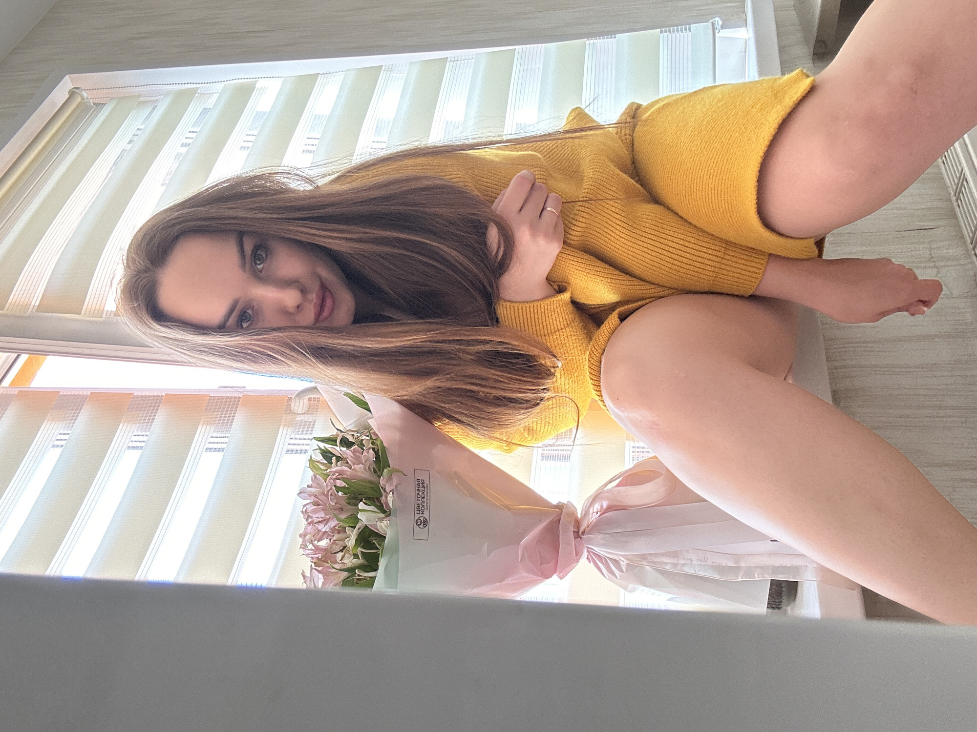 A young woman with long brown hair lies on a surface with a bouquet of pink and white flowers. She is wearing a yellow sweater. The background includes a window with blinds. The city of Chaikovsky, Russia cannot be definitively determined as the location based on the provided image alone.