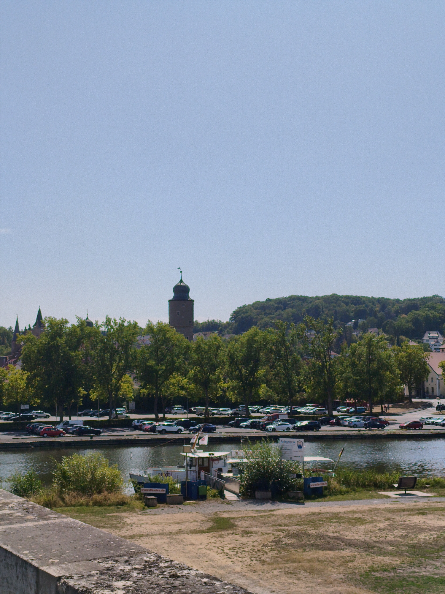 The image shows a view of the Main river in Ochsenfurt, Germany. In the background, there is a town with several buildings, including a prominent tower. The foreground features a boat docked on the riverbank, with trees and a parking lot visible behind it. The scene is bathed in sunlight, with a clear blue sky overhead. The river reflects the surrounding scenery, creating a peaceful and picturesque atmosphere.