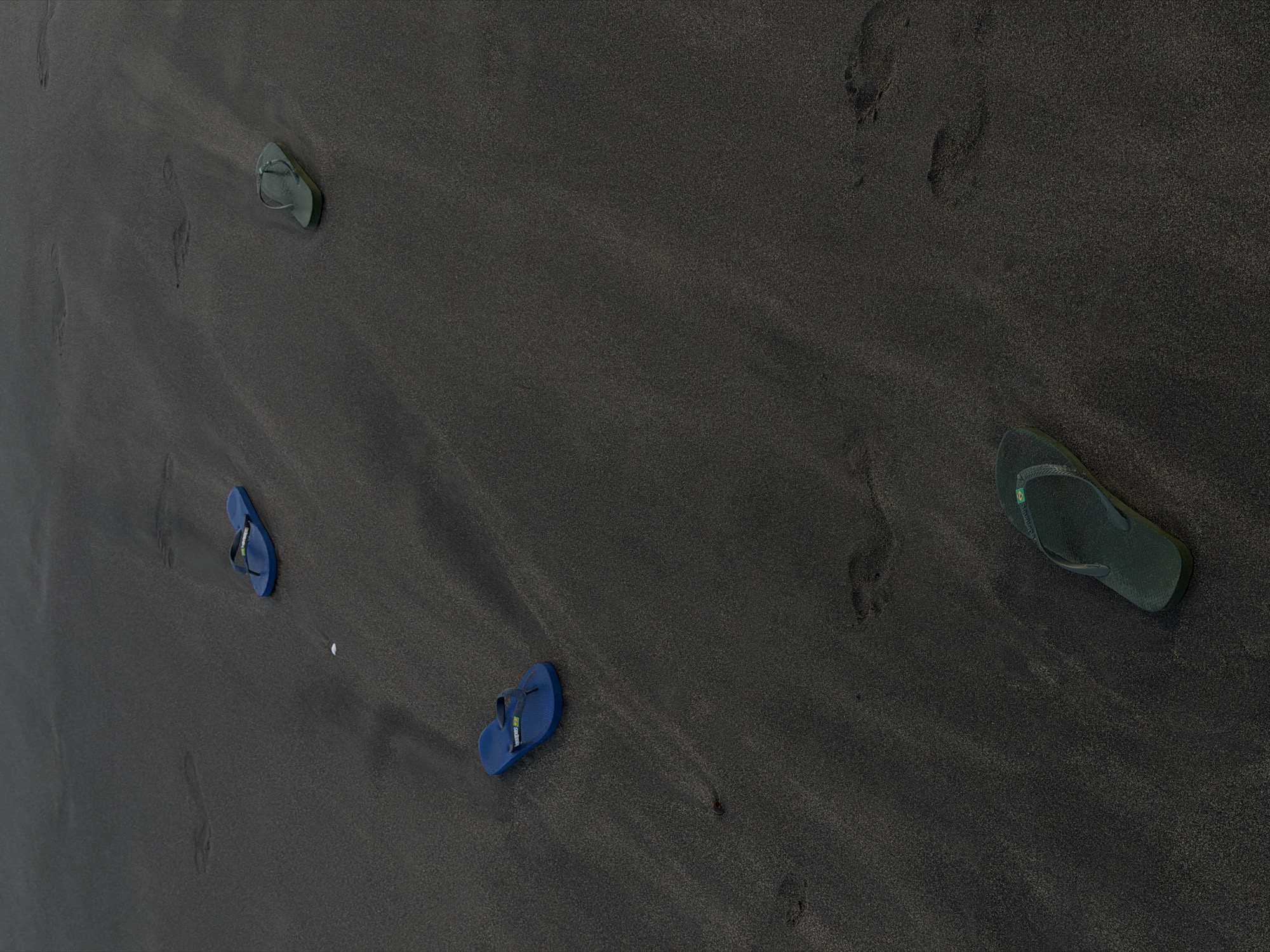 Four flip-flops are scattered on a dark, sandy beach. Two blue flip-flops are located in the upper left quadrant of the image, one near the top edge and another slightly below and to the left. Two green flip-flops are visible, one in the upper right quadrant and another near the bottom edge of the image. Footprints are visible in the sand throughout the scene. The location is identified as the city of North Kuta, Indonesia.