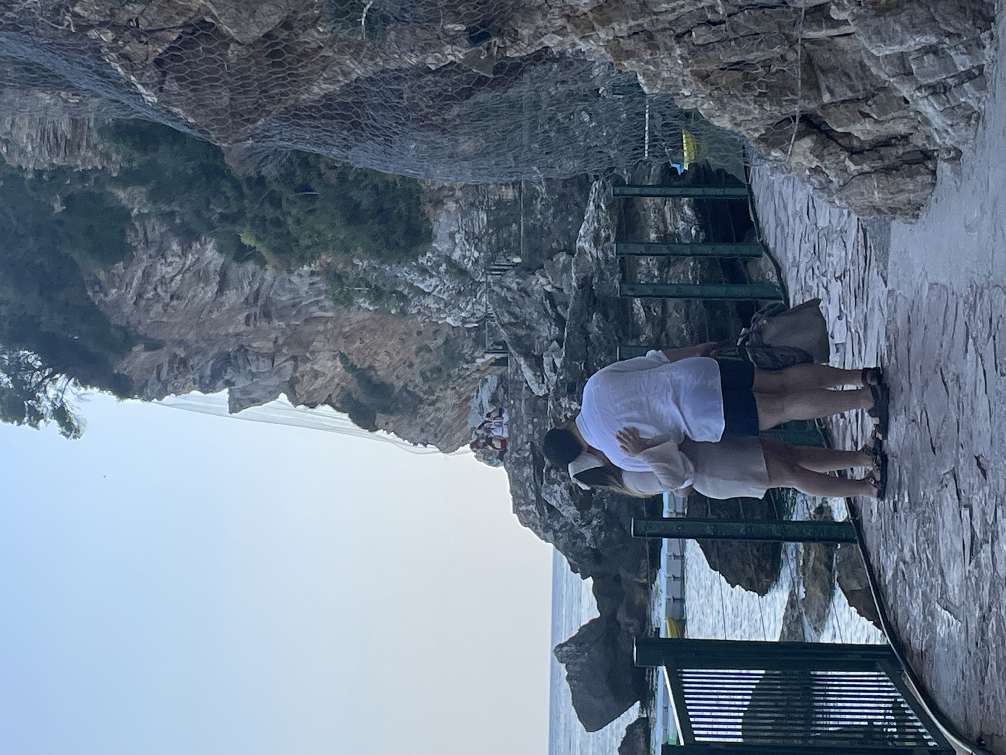 The image shows a couple embracing on a stone walkway, likely a tourist spot, built along a rocky coastline. The man wears a white shirt and dark shorts, while the woman wears a beige dress. The walkway features green metal railings and a structure resembling a cage or net covers the rock face above. A glimpse of the sea is visible below. In the background, another group of people is faintly visible. The location is likely near a rocky coast in Budva, Montenegro, known for its coastal walkways and dramatic rock formations.