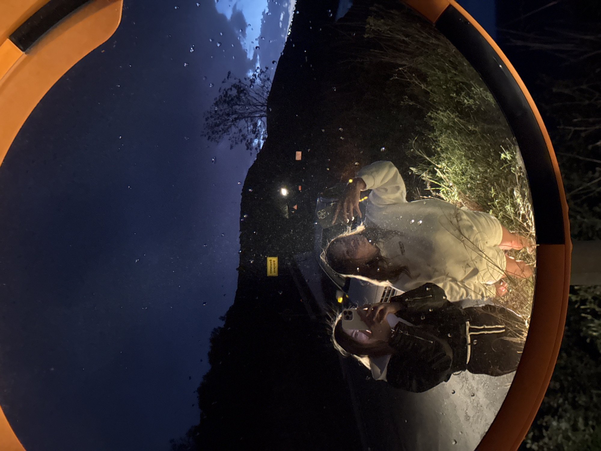 The image displays a distorted reflection within an orange and black-framed convex road mirror, captured outdoors at night in Visnjeva, Montenegro. The mirror's surface is covered with numerous water droplets, indicating recent rain or high humidity.
Two individuals are visible in the reflection. The person in the foreground wears a dark jacket, possibly black with white stripes on the sleeves, and holds a smartphone, appearing to take the picture. Their long, light-colored hair is visible around the phone. Behind them, a second person, dressed in a light-colored long-sleeved top or hoodie, is reflected in a sitting or crouching posture.
The reflected background features a dark night sky with visible cloud formations and the silhouette of a bare tree in the upper left section. Dark, sparse vegetation is reflected in the lower right, and several small, bright artificial lights punctuate the dark landscape. A small, inverted yellow sign with black text, likely Cyrillic, is also reflected near the center of the image. The entire composition is rotated vertically, presenting the sky on the left and the ground elements on the right, which adds to the surreal effect of the wide-angle reflection.