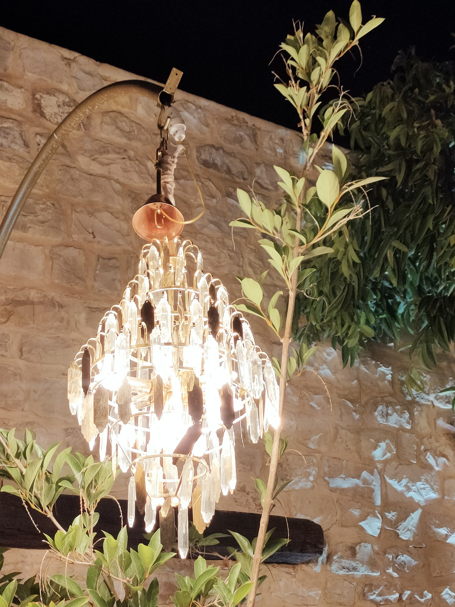 The image depicts a chandelier hanging outdoors at night, in front of a stone wall. The chandelier is ornate, with numerous clear, elongated crystal-like elements and is illuminated from within, casting a warm glow. A plant with light green leaves grows in front of the chandelier, partially obscuring it. Behind the chandelier and plant, the rough-hewn stone wall is visible, illuminated by artificial light. To the right of the plant, darker green foliage is also visible, suggesting trees or larger bushes. The sky above is dark, indicating nighttime. The scene appears to be an exterior setting, possibly a courtyard or garden area of a building. The location is identified as Cebalat, Tunisia.