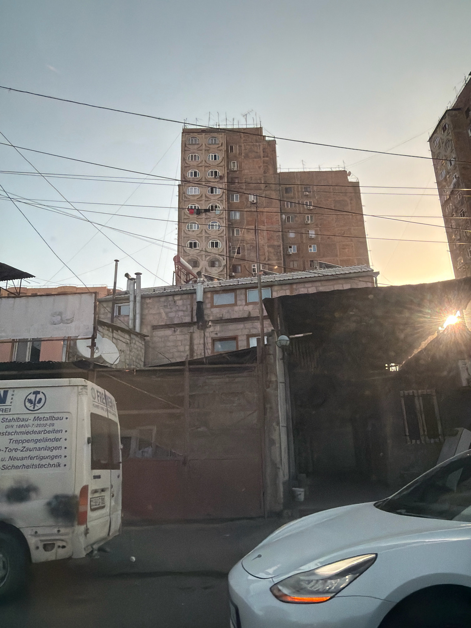 In Yerevan, Armenia, this photograph captures a cityscape with an overcast sky, suggesting either early morning or late afternoon light. The foreground features the rear of a white van, bearing text in German, indicating it's likely a commercial vehicle. To the right, the front of a white Tesla is visible, partially obstructing the view of a dark, covered structure.
In the mid-ground, a row of buildings with weathered facades lines the street, with satellite dishes and air conditioning units attached to the exteriors. In the background, towering apartment blocks dominate the skyline. One prominent building displays a distinctive architectural feature with rows of circular windows. The setting is further characterized by a dense network of overhead electrical wires, a common sight in Yerevan.
The overall impression is one of an urban environment, possibly a residential area, with a blend of older and newer construction, and the utilitarian presence of commercial vehicles. The light suggests a time of day when shadows are long, or the sun is diffused by clouds.
