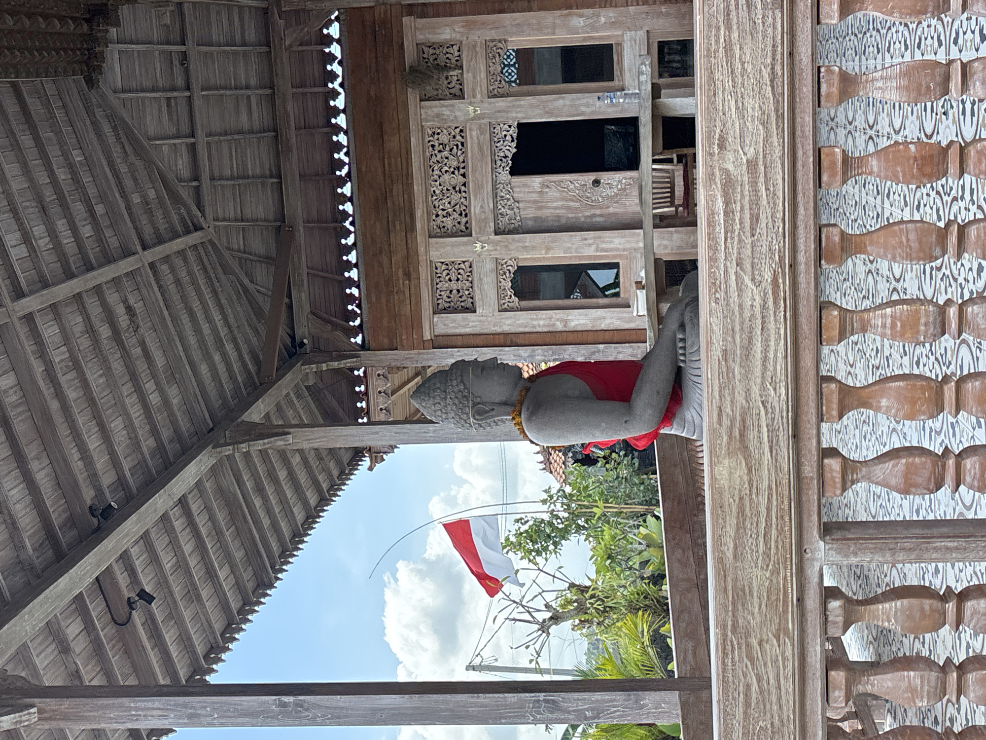 The photograph shows an outdoor scene, potentially in Kerambitan, Indonesia, featuring a statue of a Buddha figure with a red cloth draped around its neck. The statue is positioned beside a structure with intricate wooden carvings. An Indonesian flag flutters in the background, visible through an opening that frames the statue. The structure appears to be part of a building with a wooden roof, and there are decorative elements visible on the wooden exterior.