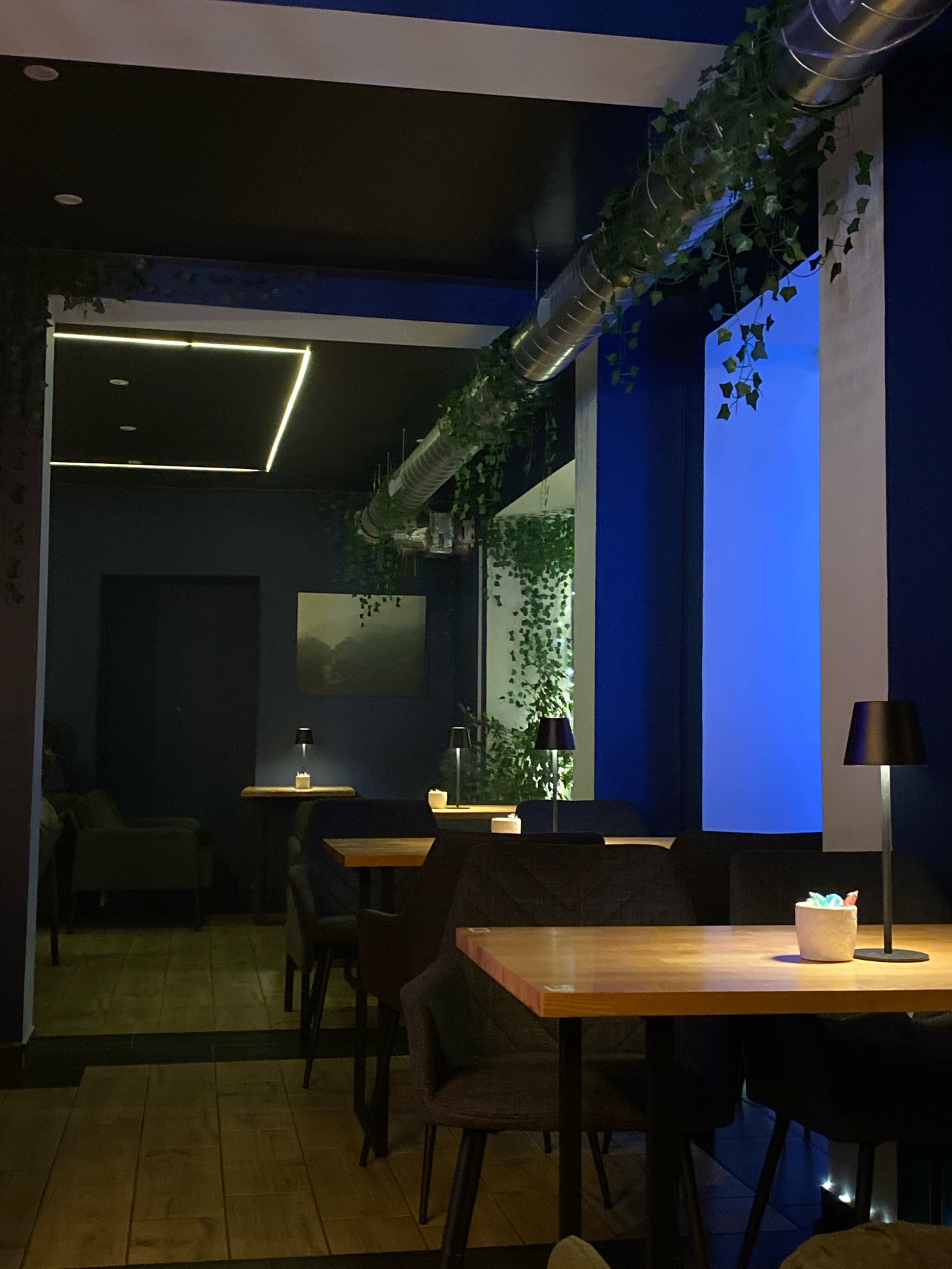 The image shows the interior of a modern cafe, possibly a lounge or a restaurant, characterized by its dark color palette, contemporary furniture, and decorative elements. The cafe is located in Gliwice, Poland.
The space is dimly lit, with ambient lighting coming from recessed ceiling lights and table lamps. A prominent feature is a large, industrial-style ventilation duct running across the ceiling, draped with artificial greenery that adds a touch of nature to the otherwise sleek design. The walls are painted in deep blues and dark shades, creating an intimate atmosphere.
Several tables and chairs are arranged throughout the space. The tables appear to be made of wood with metal legs, and the chairs are upholstered in dark fabric, providing a comfortable seating arrangement. On one of the tables in the foreground, there is a small, white container holding colorful decorative items, possibly sugar sticks or toothpicks.
In the background, a landscape painting hangs on the dark blue wall. Adjacent to it, a door leads to another part of the cafe or a private area. The floor is covered with wooden planks, laid in a way that enhances the modern aesthetic. A large vertical panel on the right side of the image emits a bright blue light, adding a dramatic visual element to the scene.
There are no people visible in the image. The scene is static and appears to be captured during a quiet period, perhaps before or after operating hours, or during off-peak times. The overall impression is one of a sophisticated and stylish establishment designed for relaxation and socializing.