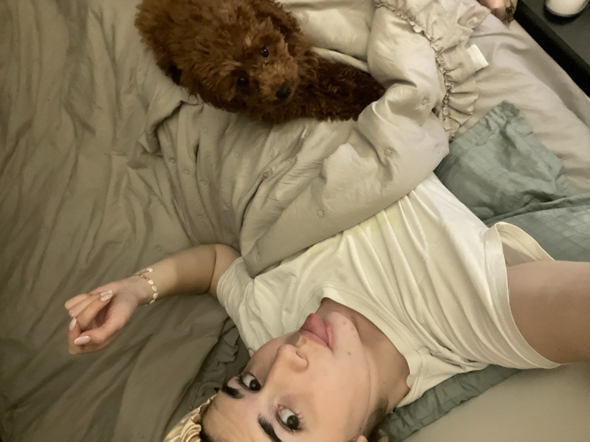 An overhead, slightly angled shot captures a person and a small dog resting together on a bed indoors. The person, appearing to be a young adult female, occupies the lower and central part of the frame, lying on her back with her head turned towards the camera. She displays a playful expression with her tongue slightly extended, has dark eyebrows, and wears a light-colored satin hair accessory around her head. Her left arm is extended to the side, revealing a white beaded or pearl-like bracelet on her wrist and light-colored manicured nails. She is dressed in a plain white t-shirt.
Above and to her left, a fluffy, reddish-brown dog, resembling a poodle, is comfortably settled on the light-colored bedding. The dog is looking directly at the camera with dark eyes, appearing calm.
The bed is covered with light grey or beige bedding, including sheets and a comforter, with some sections featuring decorative ruffles and others in a light green or teal shade. The lighting in the scene is dim, suggesting an indoor setting during the evening or night. A dark surface, possibly a bedside table, with a white cylindrical object on it, is partially visible in the upper right corner of the frame. The overall atmosphere conveys a relaxed, casual, and intimate moment, likely a selfie with a beloved pet. There are no discernible visual cues within the image itself to confirm the specified location of Sofia, Bulgaria. No visible text is present.