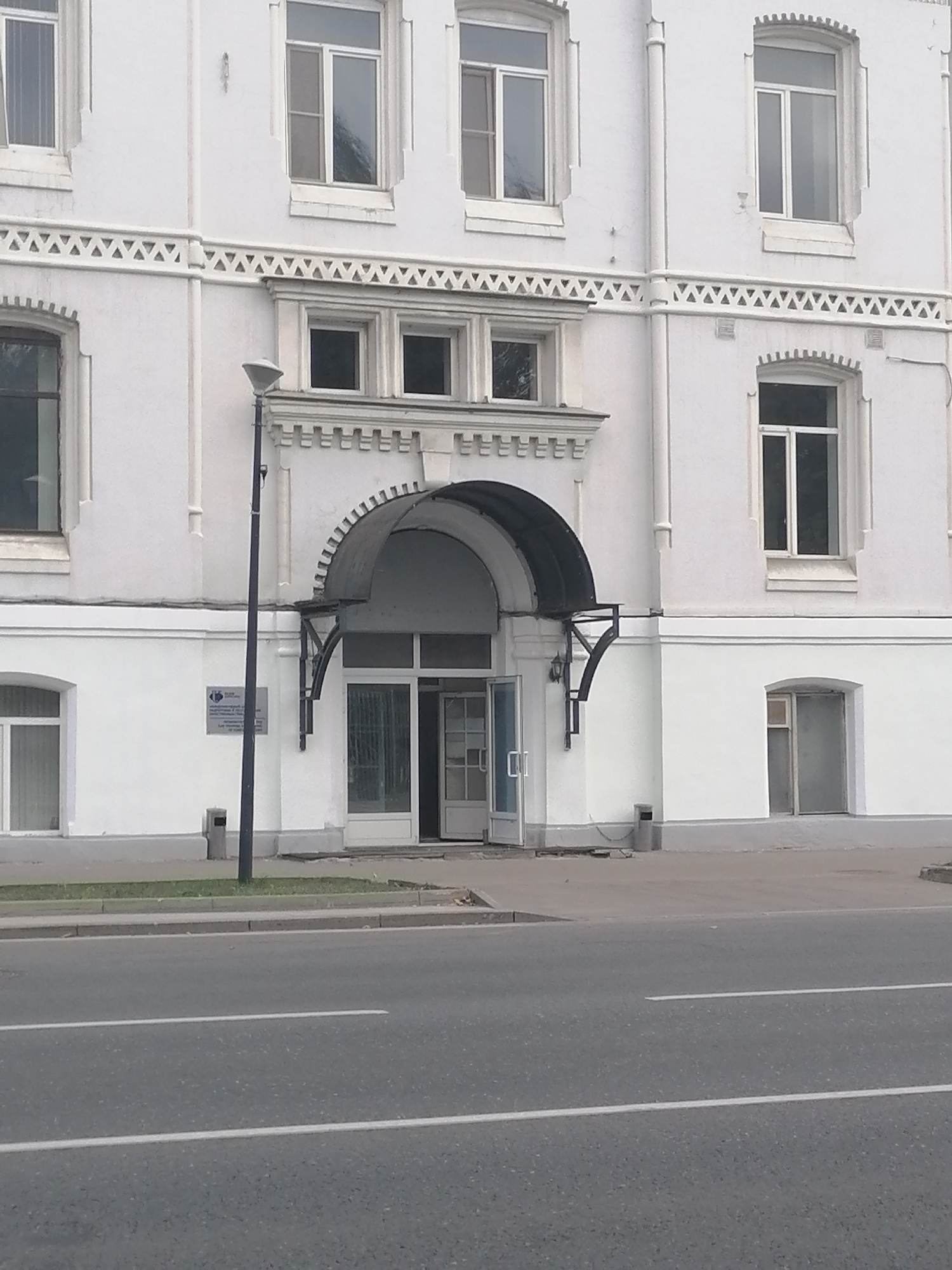 The image shows a street-level view of a white, multi-story building in Moscow, Russia. The building has a classical architectural style with decorative elements above the windows. There are several windows visible on the facade, some of which are large and rectangular, while others are smaller. An arched entrance with a dark awning leads into the building. A street lamp is positioned to the left of the entrance. A sign with text is mounted on the wall to the left of the entrance. A trash receptacle is visible near the building. The building is situated next to a road with lane markings, suggesting it is on a street in a developed urban area. The sky is overcast.
