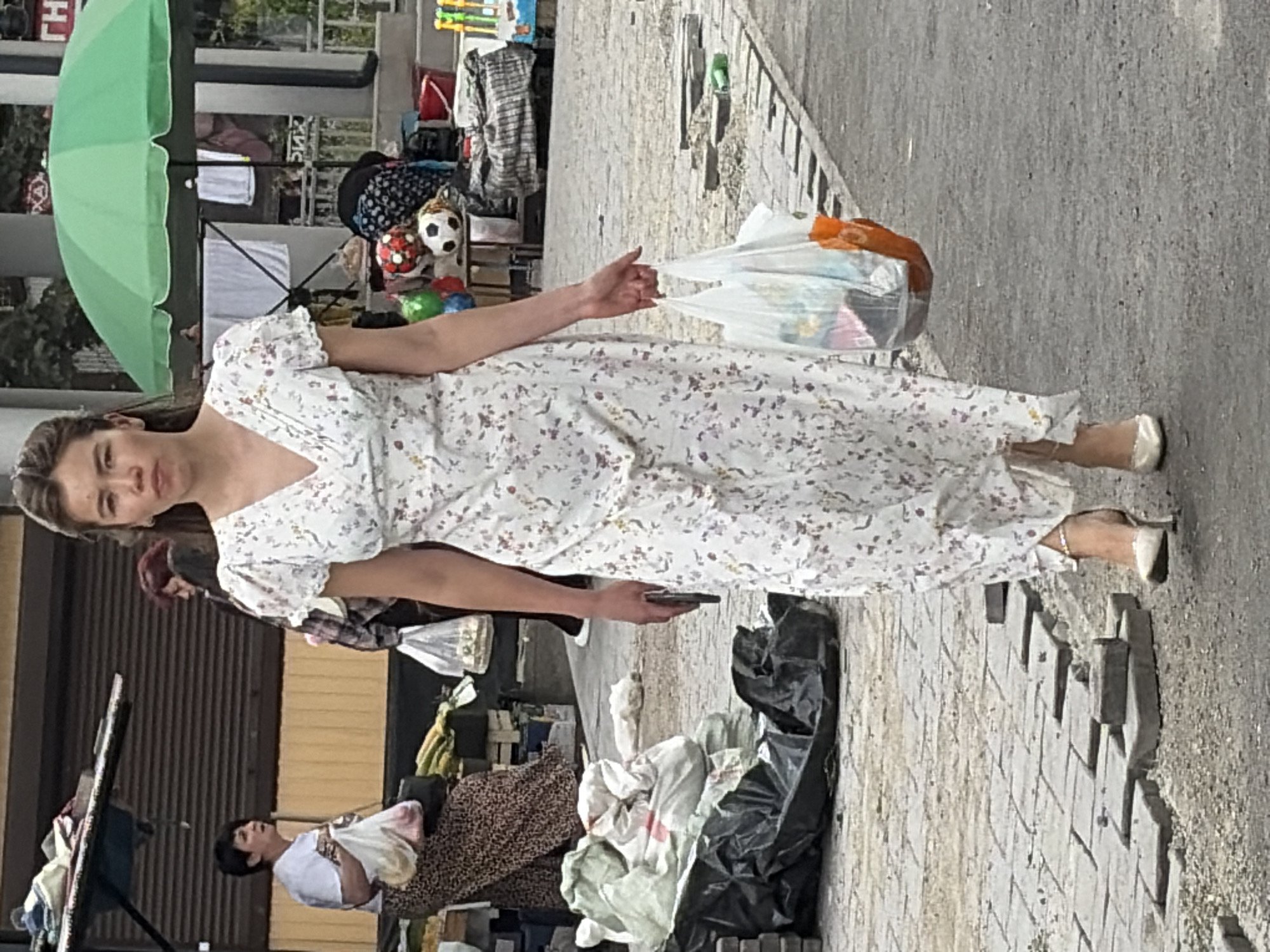 A woman in a white floral dress walks down a cobblestone street in what appears to be an outdoor market. She is holding two plastic bags filled with items, one in her right hand and another in her left. Her left hand is also holding a smartphone. In the background, other people are visible, as well as market stalls with various goods, including produce, clothing, and toys. A green umbrella is set up over one of the stalls. The ground is paved with cobblestones, and some areas appear to be under repair. The overall environment suggests a busy public space, possibly in a city.
