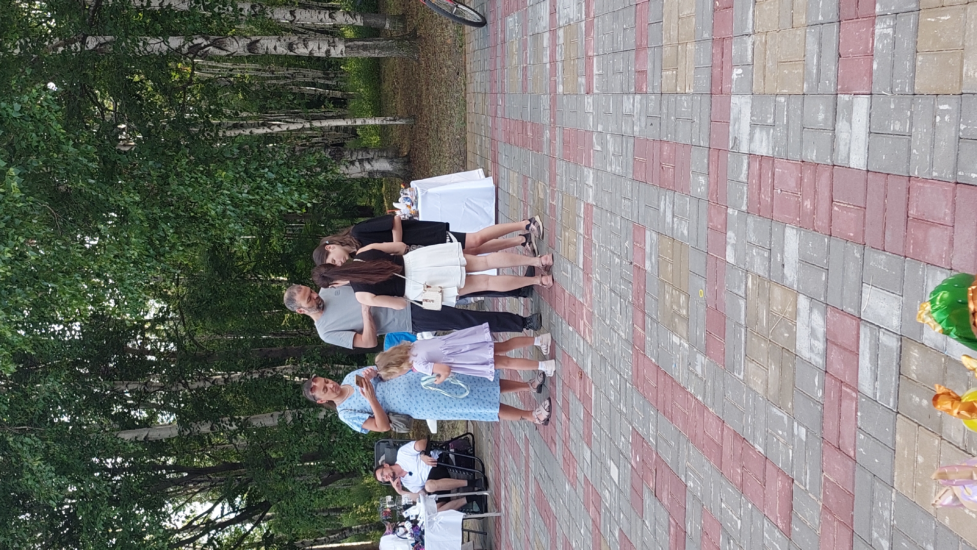 The image depicts an outdoor gathering in a park setting, likely in Komsomolsk-on-Amur, Russia, characterized by mature trees with birch and possibly other deciduous species. The ground is paved with interlocking bricks in a pattern of red, gray, and beige. In the mid-ground, a group of people are interacting. A woman in a light blue dress is looking at her phone, standing next to a young girl in a lavender dress. A man in a gray t-shirt is also looking at a phone, facing two teenage girls. One of the girls is wearing a black sleeveless top and a white skirt, and the other is wearing a black t-shirt. Behind them, a white tablecloth is set up, possibly for food or drinks, with some items visible on it. To the left, a man is seated in a black folding chair at a table with a white tablecloth, wearing a white t-shirt and dark shorts. Further to the right, part of a bicycle wheel is visible. In the foreground, decorative balloons are partially visible on the paving stones. The overall scene suggests a casual event or picnic in a park.