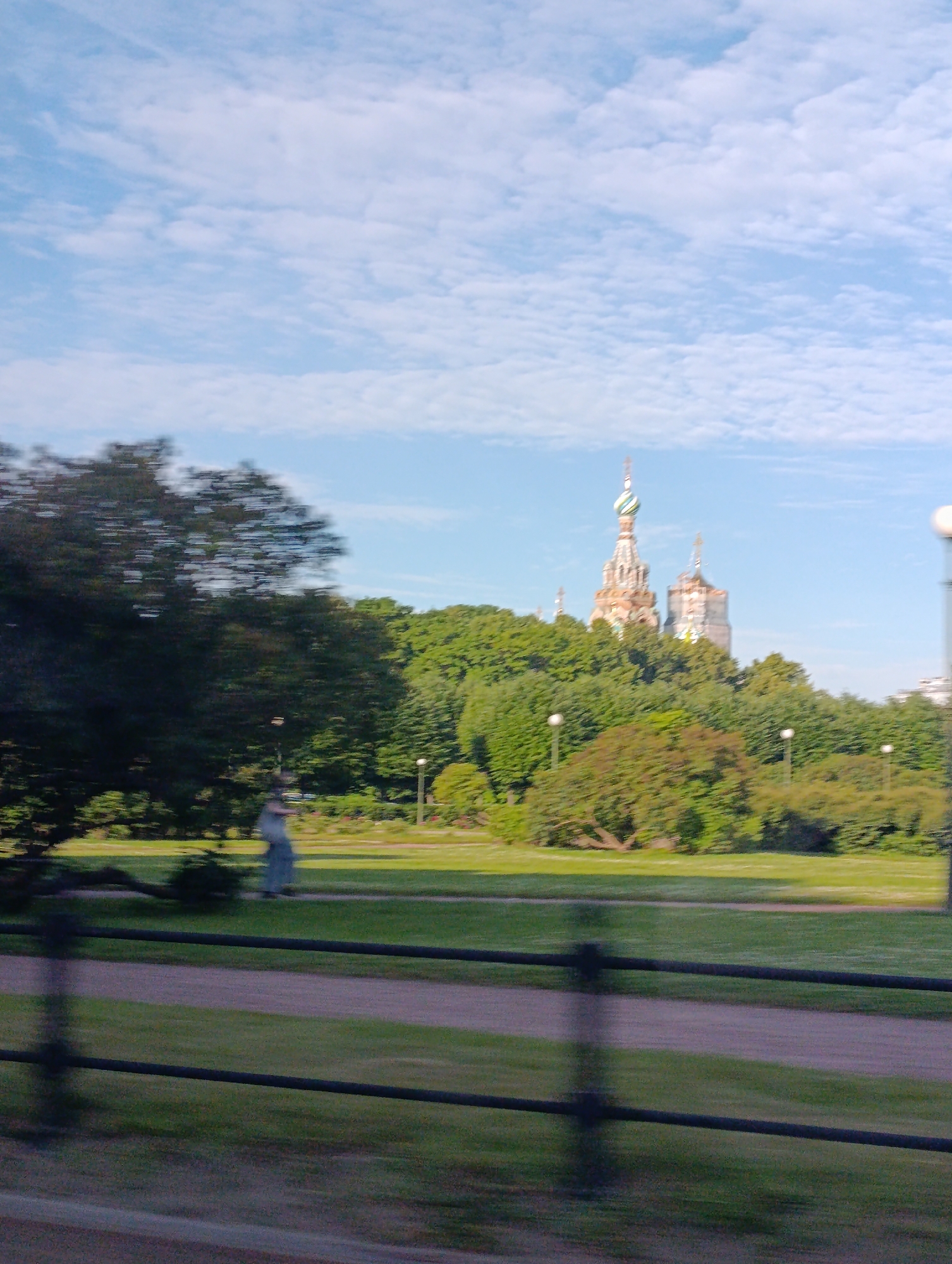 The image depicts a view of the Savior on Spilled Blood church in Saint Petersburg, Russia, from a distance. The church's distinctive onion domes and intricate facade are visible above a dense canopy of green trees. In the foreground, a blurred fence separates a grassy area with a path from a darker area that appears to be a road or another path. A solitary figure, also blurred, is seen walking on the grass in the mid-ground. The sky is a pale blue with scattered white clouds. The overall impression is of a bright, possibly sunny day, with the motion blur suggesting the photo was taken from a moving vehicle.