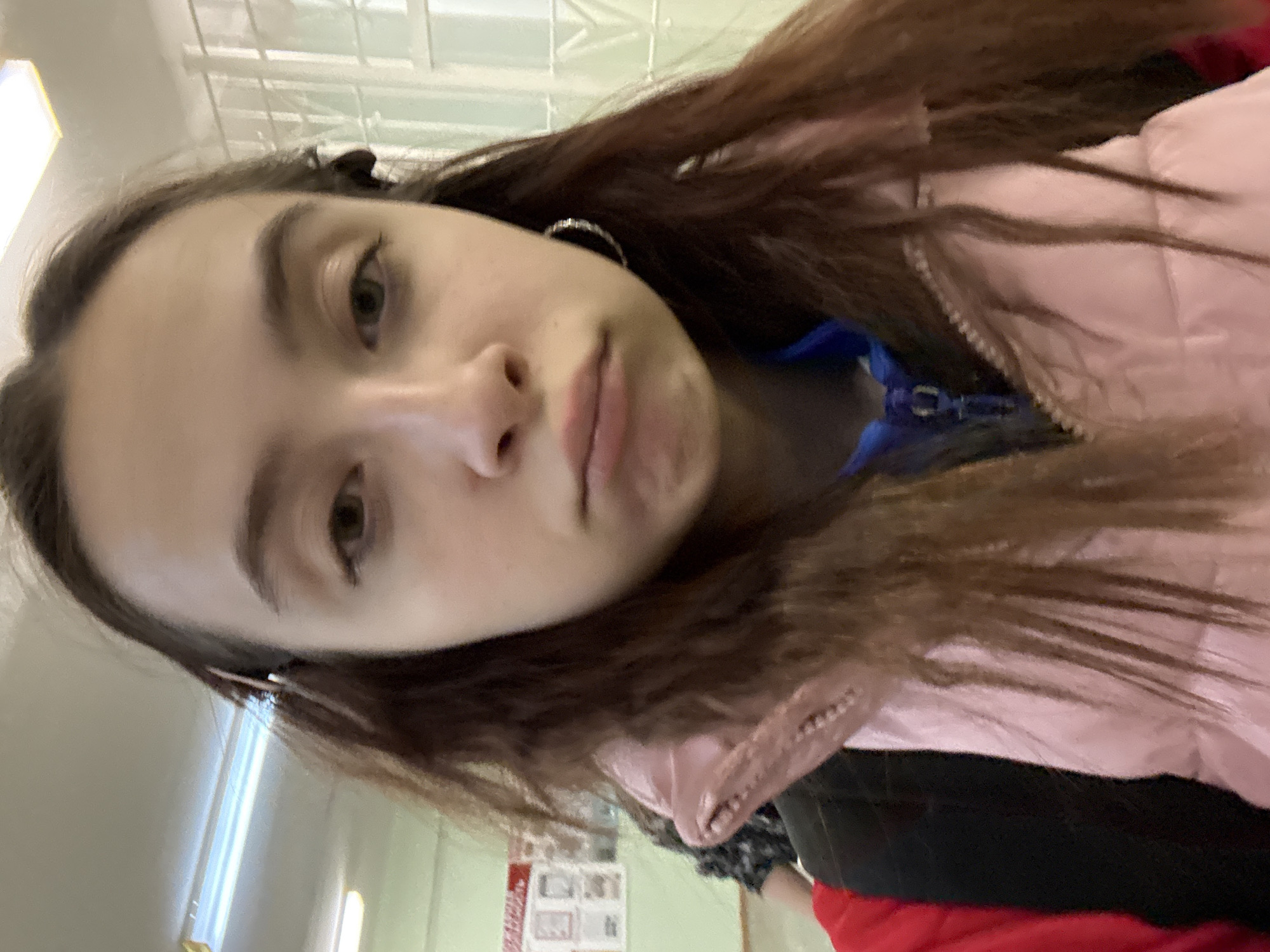 A selfie of a young woman with long brown hair, wearing a pink jacket and blue shirt. She is indoors, as evidenced by the light fixture and wall behind her. In the background, there is a notice board with papers on it, and a window. The location is Berezniki, Russia.