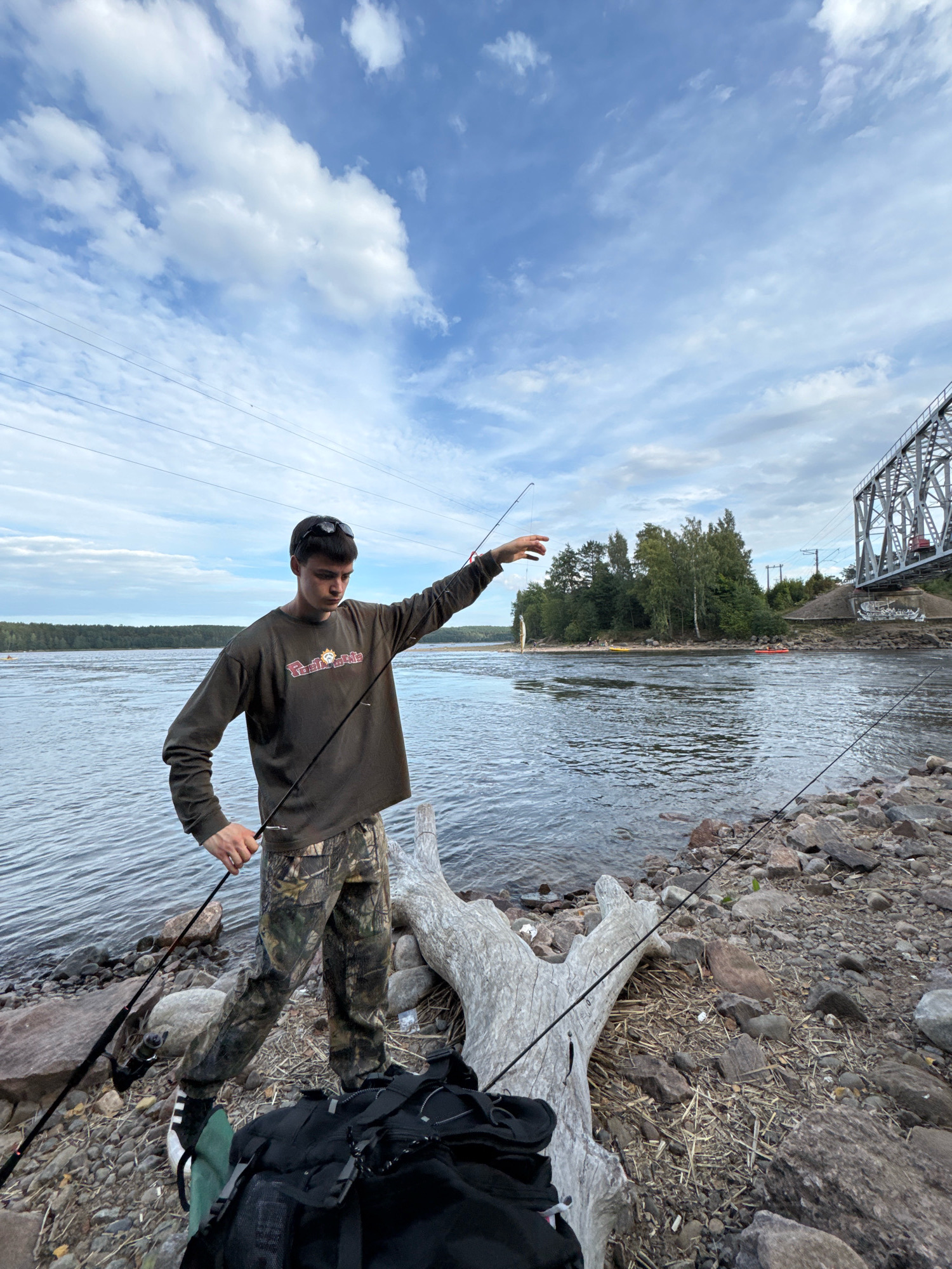 The image depicts a young male, appearing to be in his late teens or early twenties, standing on a rocky shoreline next to a body of water. He is wearing a long-sleeved olive-green shirt with a graphic print and camouflage-patterned pants. He holds a fishing rod in his right hand, which extends towards the water, and his left hand is near the reel. Behind him, a large river flows, and beyond that, a tree line borders the opposite bank. In the background, a large metal railway bridge spans the river. Two kayaks, one yellow and one red, are visible in the water near the far bank. A large piece of driftwood lies on the shore near the subject, and a black backpack is positioned at his feet. The sky is partly cloudy with a mix of white clouds and blue sky. The scene suggests a recreational fishing activity by the river, with the railway bridge indicating a developed area, possibly Losevo, Russia, given its known railway infrastructure and river access.