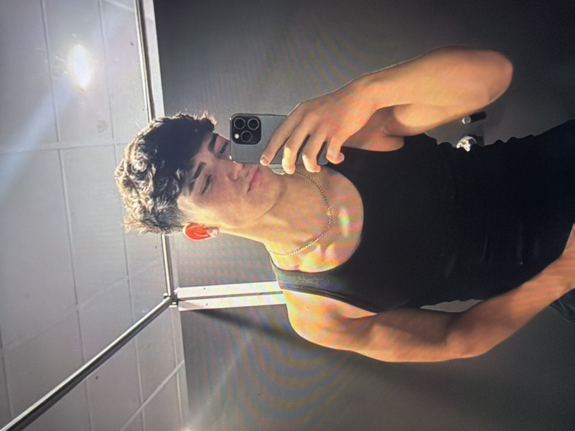 In Shumen, Bulgaria, a young male is depicted in an indoor setting, taking a mirror selfie. The subject, appearing to be Caucasian, has dark, curly hair and is wearing a black tank top and a gold-colored chain necklace. An earring is visible in his left ear. He holds a dark-colored smartphone, equipped with a multi-lens camera array, in his right hand to capture the image. His left arm is partially visible and appears flexed. The background features a white tiled ceiling with an illuminated circular light fixture, a horizontal metal bar positioned at the top left, and dark-toned walls. Vertical white structural elements are visible behind the subject's right side. The scene is illuminated by overhead lighting, creating reflections and highlights on surfaces and the subject's skin.