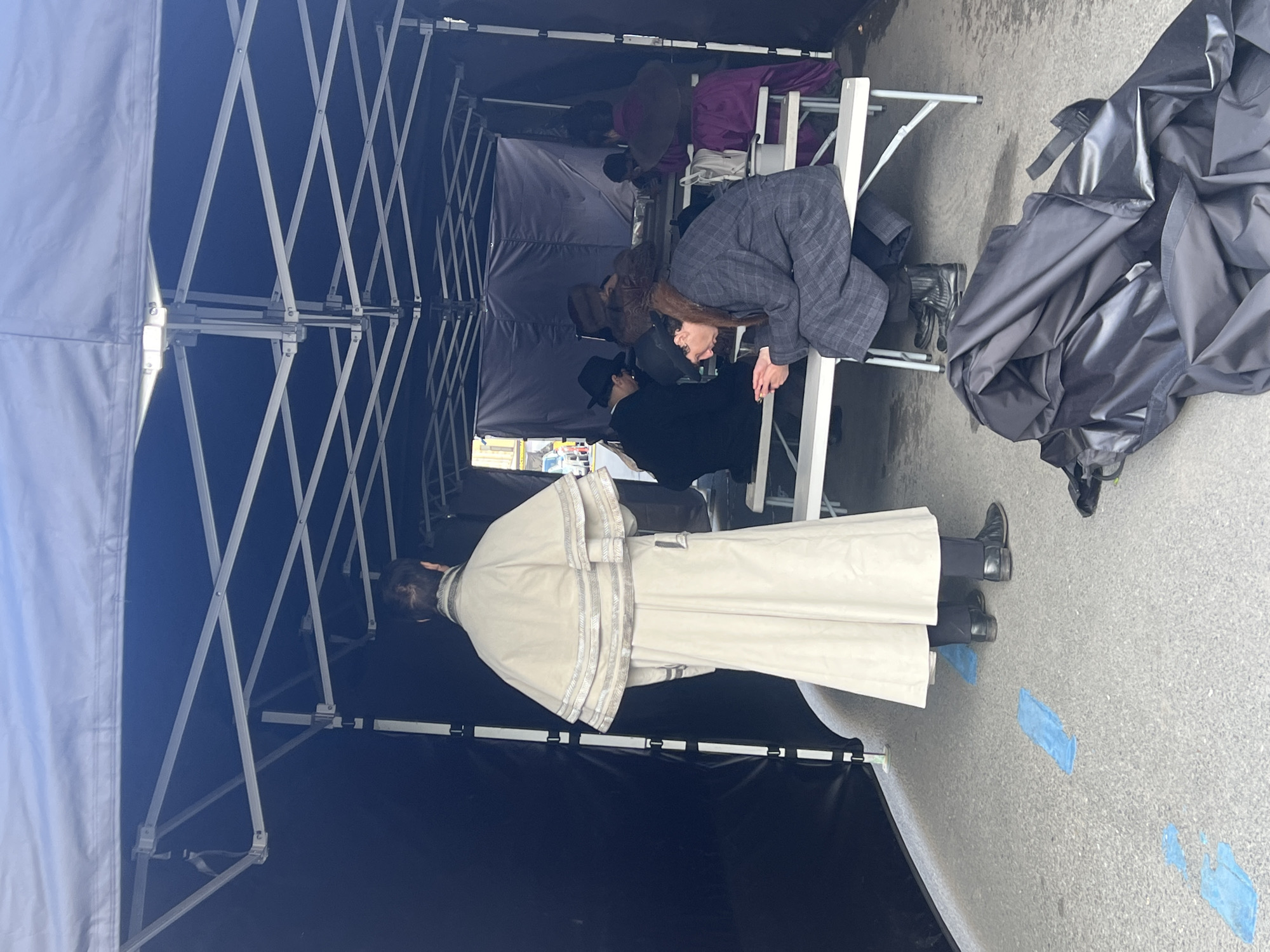 The image depicts a film set within a blue pop-up tent, presumably in Saint Petersburg, Russia, given the clothing styles and presumed filming location. Inside the tent, individuals are preparing for a scene. One person wears a long, cream-colored coat with silver trim, and a hat. Another is in a grey checkered suit. The tent is situated on a paved surface with blue tape marking positions. A pile of black material or tarpaulin is present near the tent's edge. Additional people are present in the background, but their roles are unclear. The scene suggests a production behind-the-scenes setup.