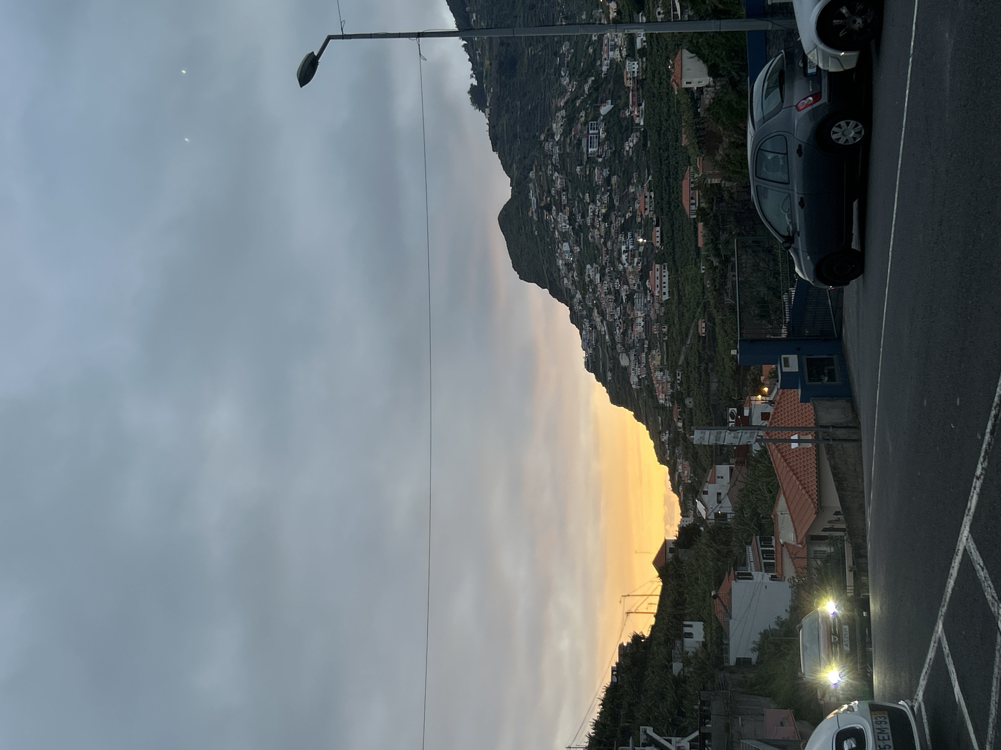 The image depicts a view in Câmara de Lobos, Portugal, during twilight. A mountain slopes down to a town with densely packed houses, their red-tiled roofs visible. The sky is cloudy, with the lower portion tinted orange from the setting sun. A street runs along the right side of the image with parked cars. A streetlight is present on the left.