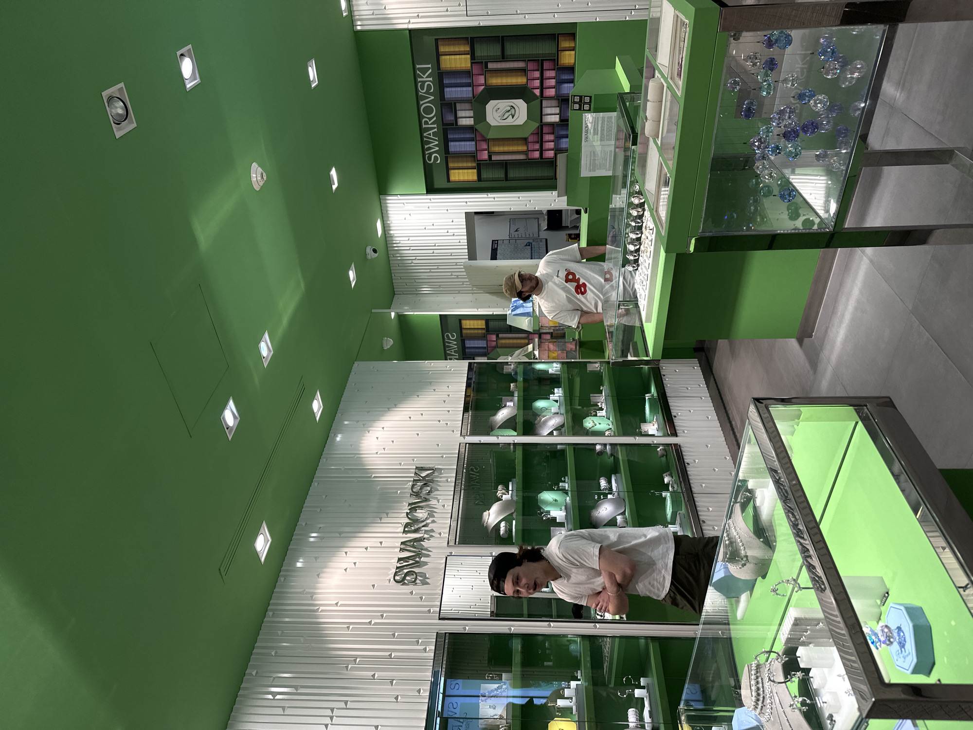 A sideways-oriented indoor shot shows the interior of a Swarovski store, presumably in Huixquilucan de Degollado, Mexico. Two people are visible: one wearing a white t-shirt, khaki pants, and a black beanie is standing at a glass display case filled with jewelry, his arms crossed; another person is seen near the counter wearing a white t-shirt with a red design and a beige hat. The store is decorated with green walls and white accent panels. The Swarovski logo is displayed prominently on the walls. Jewelry is showcased in multiple glass display cases. The lighting is provided by recessed square spotlights.