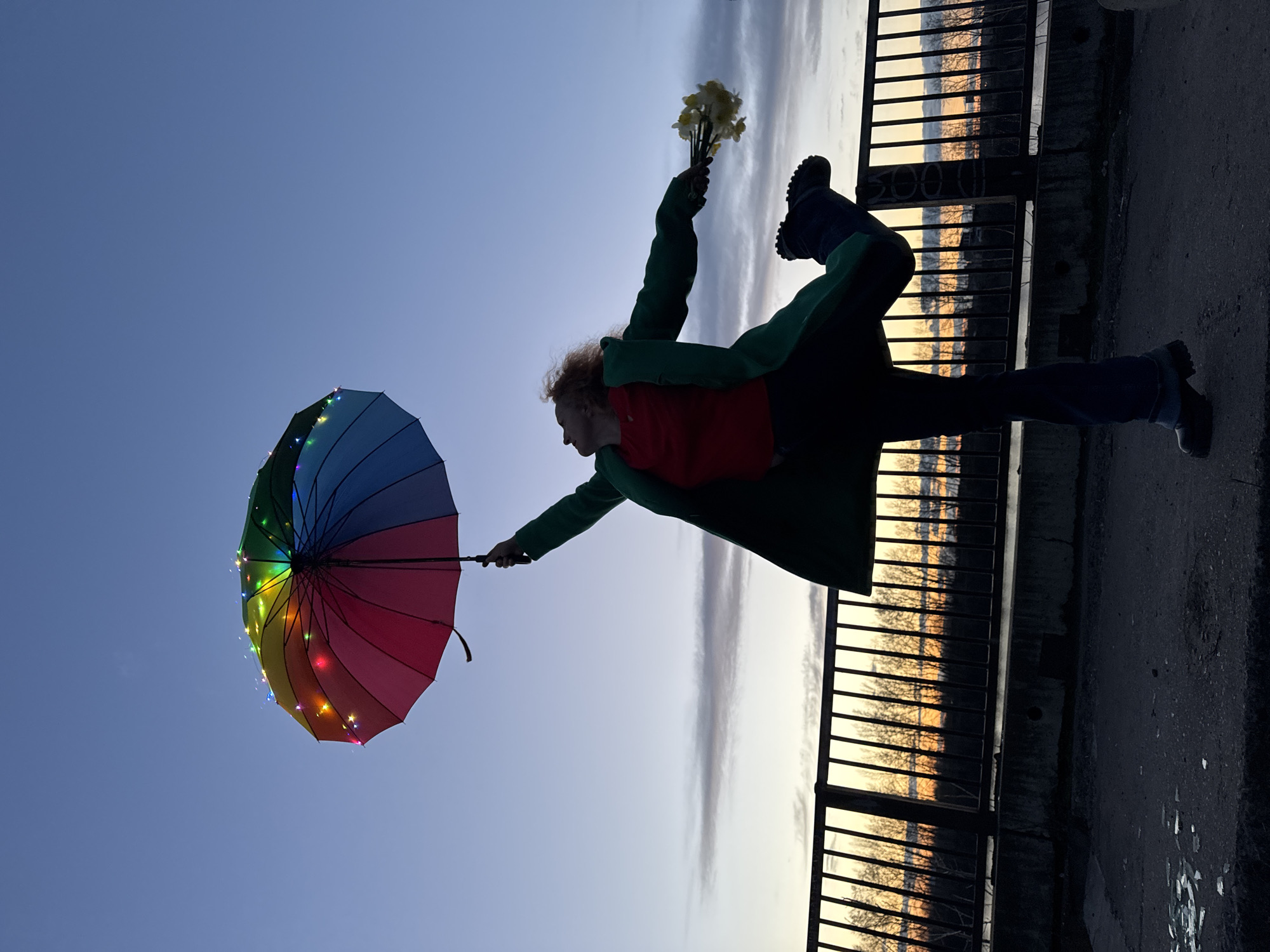 An outdoor, full-body shot captures a person in a dynamic, artistic pose against a twilight sky in Chaikovsky, Russia. The individual, seen in near-silhouette, has reddish-brown curly hair and is wearing a green jacket over a red garment, with dark trousers and sturdy boots. They are balanced on their right leg, with the left leg bent upwards behind them, creating a sense of movement or dance.
In their left hand, the person holds a vibrant, rainbow-colored umbrella adorned with small, multi-colored LED lights that glow softly. In their raised right hand, they hold a small bouquet of yellow flowers, possibly daffodils.
The setting appears to be an elevated position, like a bridge or embankment, overlooking a distant landscape. A dark metal fence or railing runs horizontally across the right side of the frame, separating the foreground from the background. Beyond the fence, the horizon is bathed in the warm glow of either sunrise or sunset, casting reflections on what appears to be water or a low-lying area. Bare trees are visible against the glowing sky. The sky above is a gradient of soft blue and purple hues, with wispy clouds. The ground or pavement where the person stands is dark and textured. The overall mood is whimsical and joyful, highlighted by the illuminated umbrella and the playful pose at the golden hour.