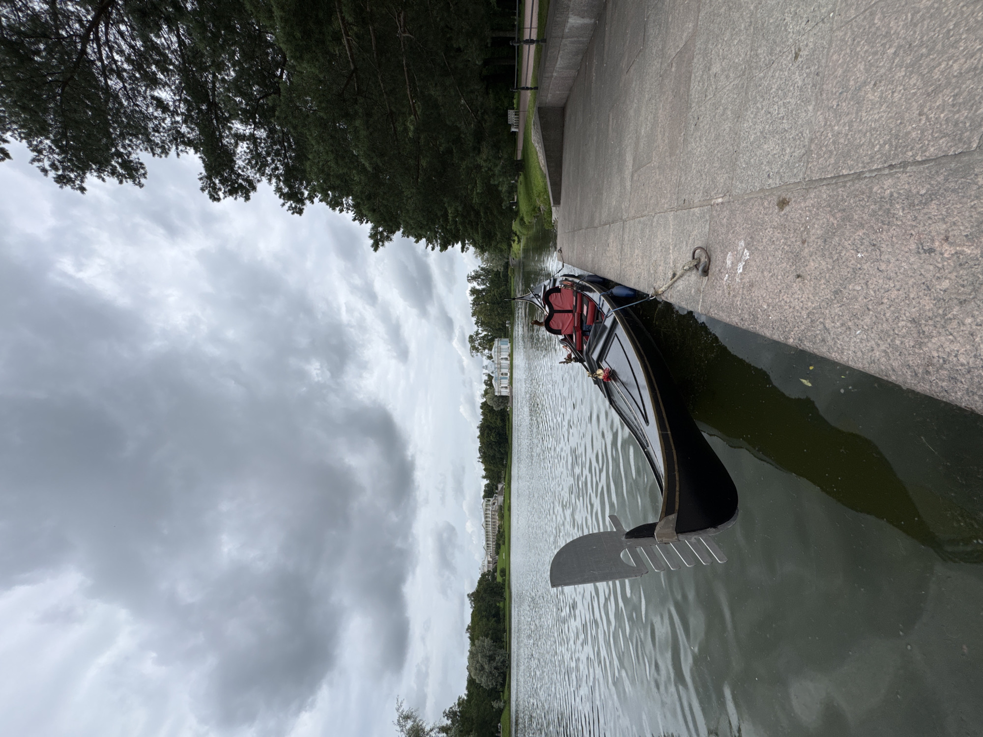 A gondola is moored to a stone embankment on a body of water, likely a canal or lake. The boat is black with red upholstery. In the background, across the water, a classical white building with a long colonnade stands on a grassy hill, surrounded by trees. To the right of the embankment, a path lined with trees is visible. The sky is overcast with heavy, grey clouds. The image was taken in the city of Pushkin, Russia, likely in the Catherine Park, given the architecture and landscape.