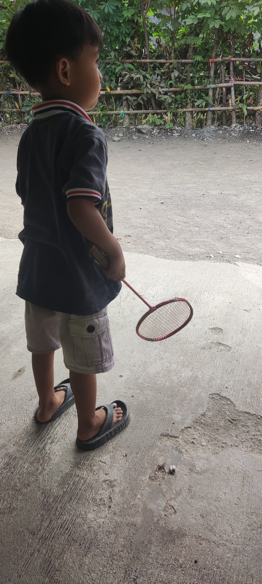 A young child stands on a concrete surface in Karangwangun, Indonesia, facing away from the camera and to the right. The child is wearing a dark t-shirt with red and white trim on the collar and sleeves, and light-colored cargo shorts. The child's feet are clad in dark gray flip-flops. In their right hand, the child holds a red badminton racket, with the head of the racket angled downwards. The background consists of a bamboo fence, behind which foliage is visible. The ground is a rough, textured concrete area with some scattered debris.